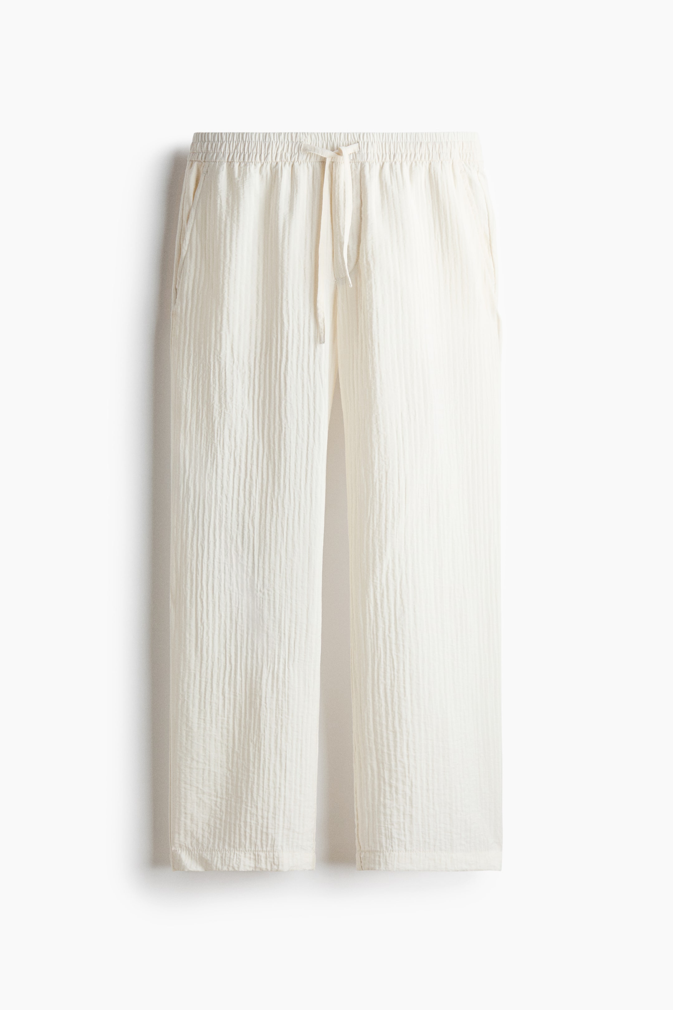 View larger image: The off-white pants are displaying a crinkled texture, an elasticated waistband with a drawstring, and side pockets. They are presenting a loose, wide-leg fit from the front.