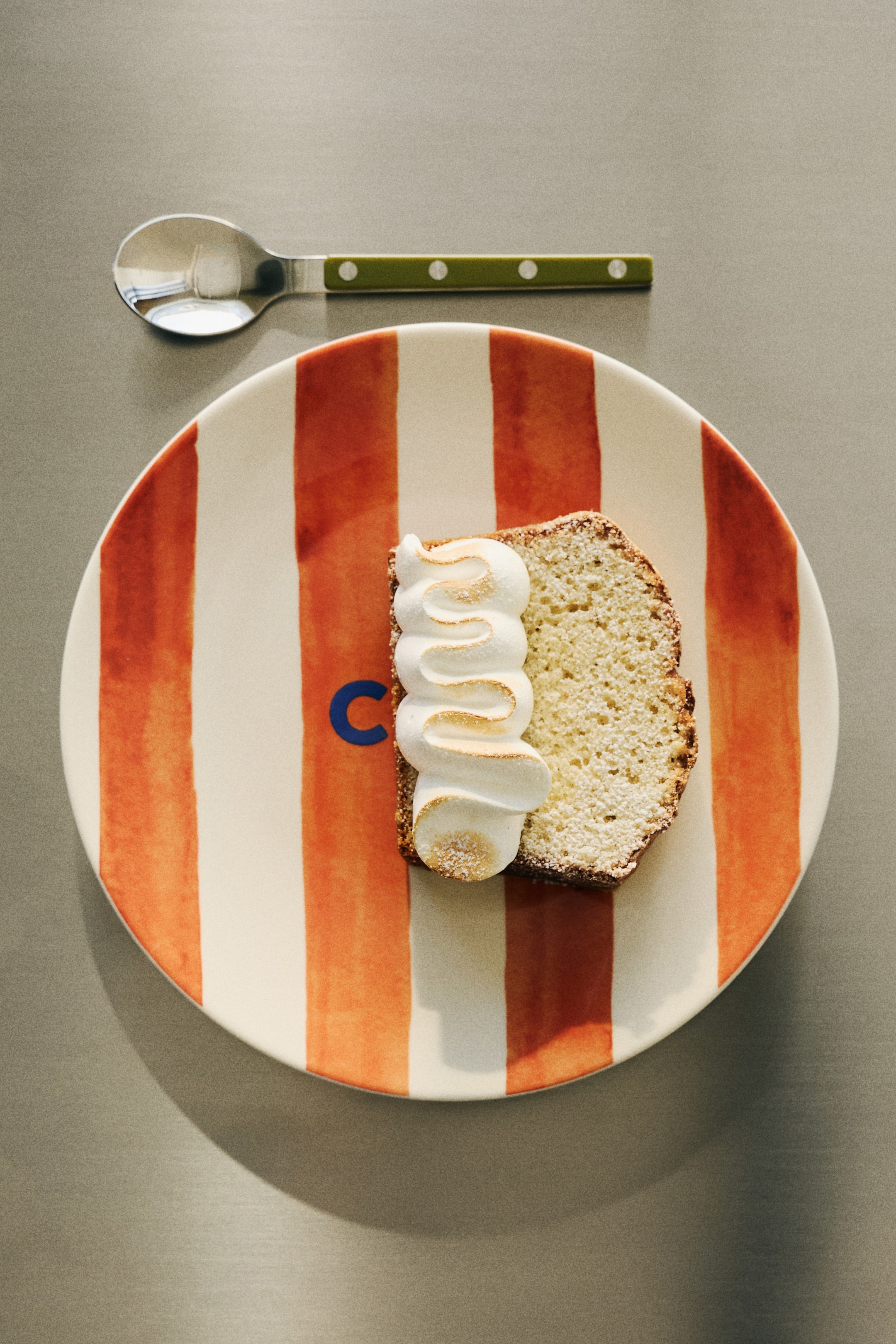 Agrandir l'image: Une tranche de gâteau, légèrement saupoudrée de sucre glace, repose sur une assiette ronde avec des rayures verticales brun-rouge et blanches. Le gâteau est surmonté d'une meringue ondulée, légèrement dorée, et un logo bleu "C" est visible sur une rayure blanche à côté du gâteau.