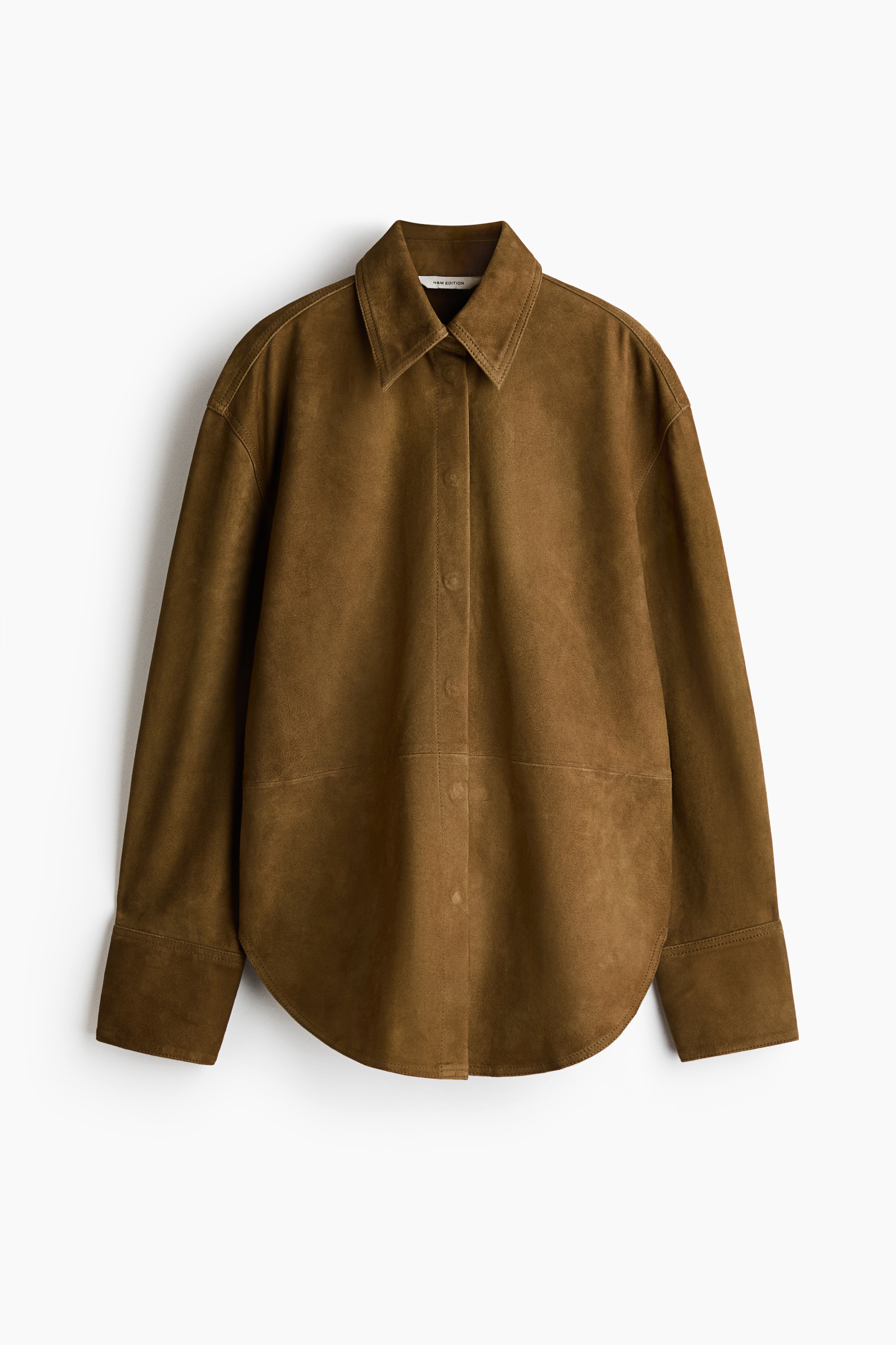 The model is wearing an oversized olive-brown suede shirt, styled open at the collar to reveal a silver press stud. The shirt is layered over a dark brown ribbed roll neck and tucked into matching trousers secured with a black belt featuring a large, ornate silver buckle.