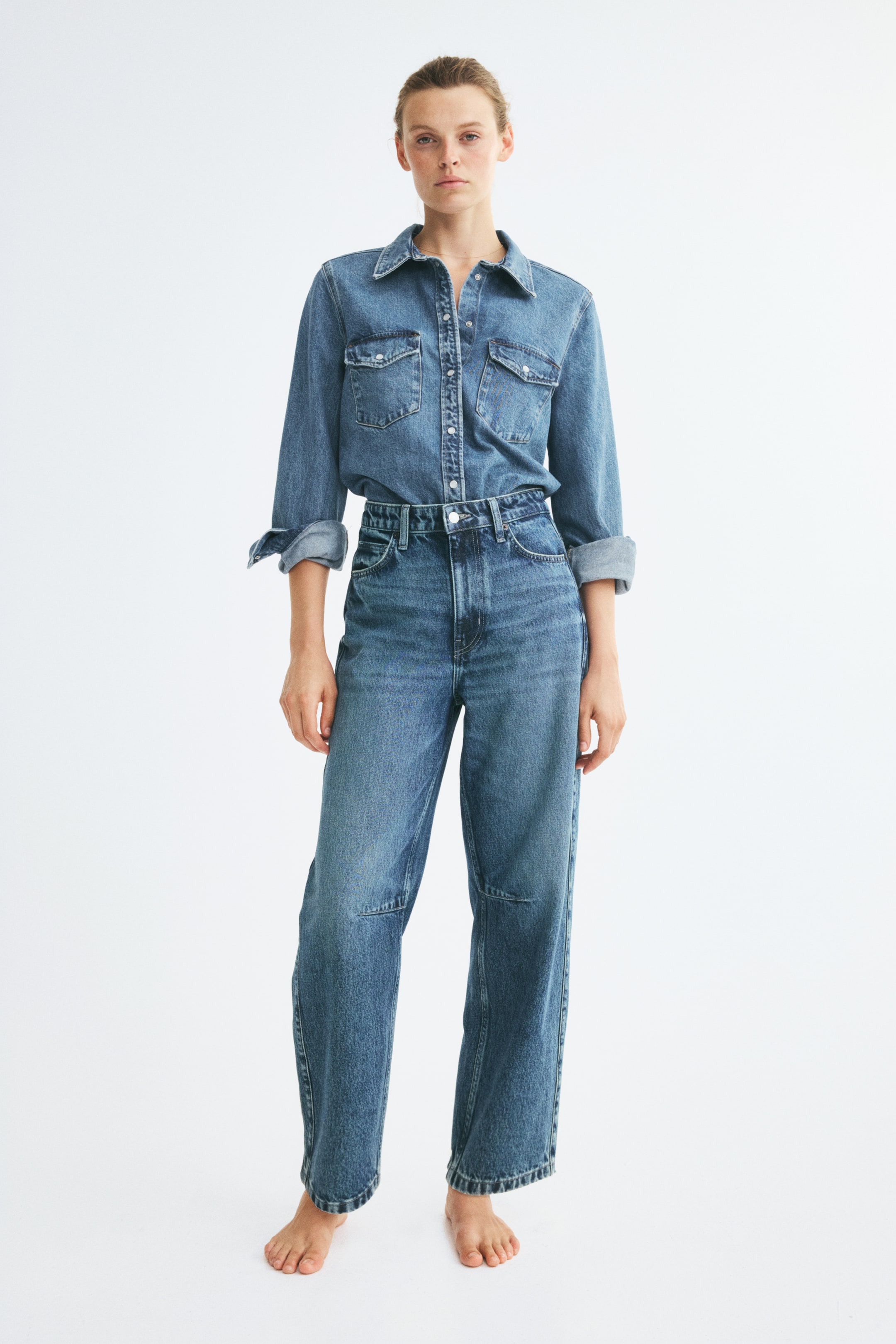 Vis større bilde: Høyt liv, blå denimjeans med buede, voluminøse ben, synlige knappedarter ved knærne og sidesømmer som vender fremover, bæres av modellen. Jeansene er stylet med en matchende denimskjorte stukket nedi linningen.
