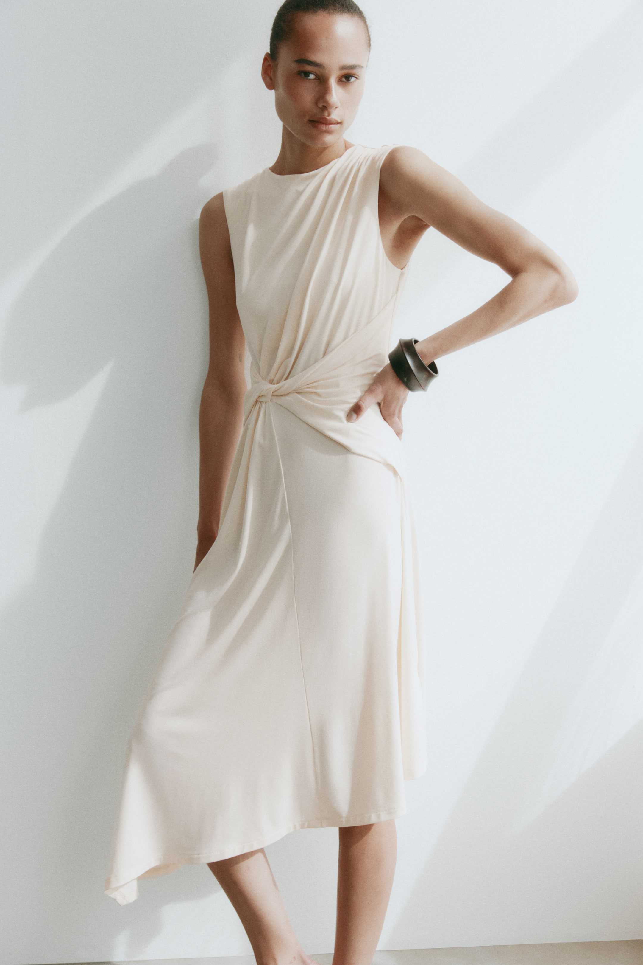 View larger image: A light cream sleeveless dress features a round neckline and a knot detail at the left waist, creating soft drapes. The fabric flows into a midi-length skirt with an asymmetrical hemline.