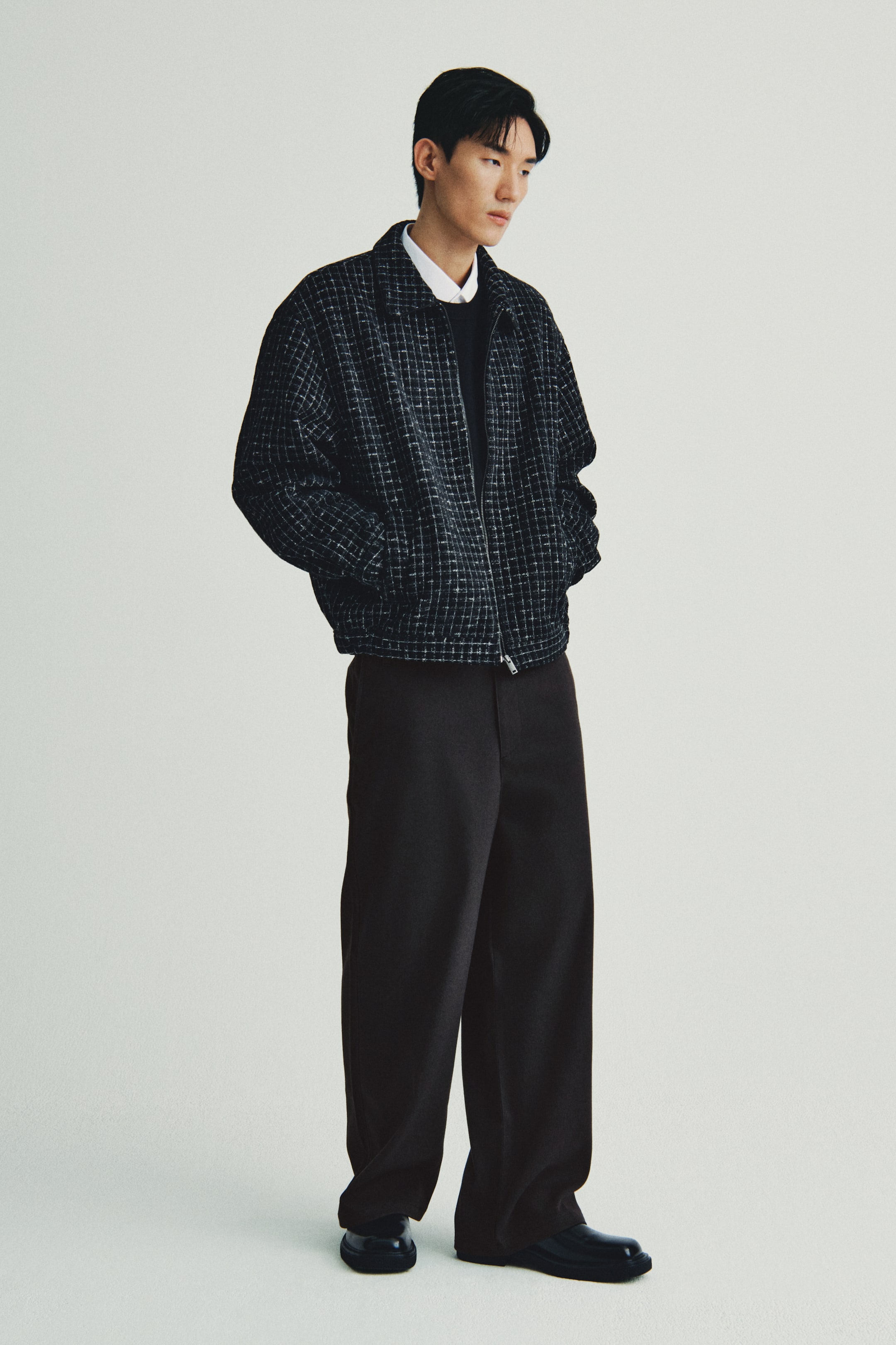 이미지 확대 보기: 어두운 회색 체크무늬 폭격기 재킷은 루즈한 핏, 풀집 클로저, 칼라, 사이드 포켓을 특징으로 하며, 약간 각도가 있는 앞면 뷰에서 흰색 칼라 셔츠와 검은색 크루넥 톱 위에 착용하고 있습니다. 검은색 와이드 레그 바지와 검은색 광택 나는 청키솔 신발과 매치되어 있습니다.