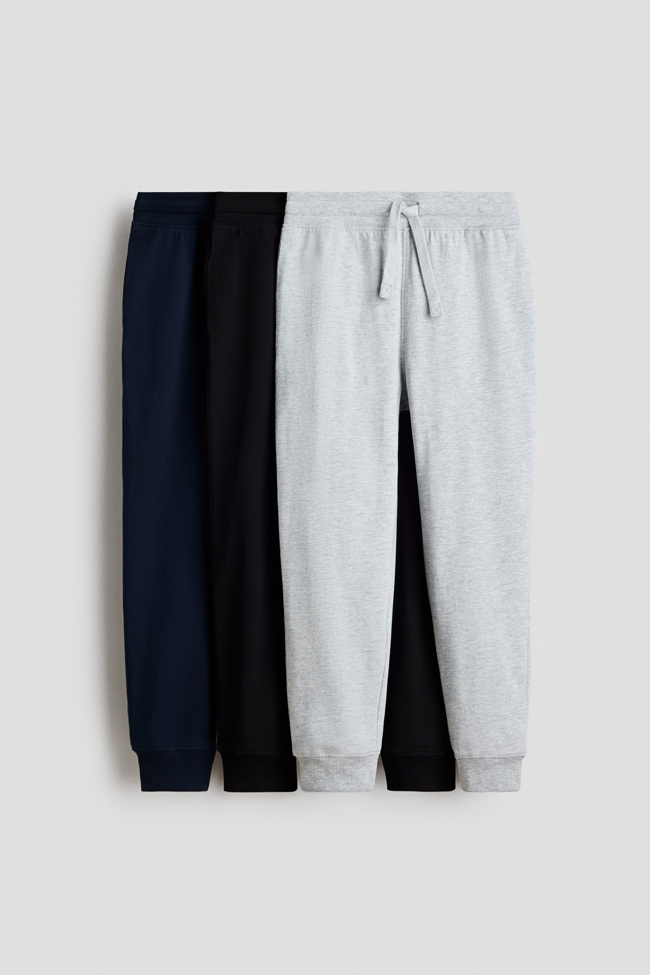 Agrandir l'image: Trois paires de pantalons de survêtement sont présentées : un bleu marine, un noir et un gris clair chiné. Les trois paires possèdent une taille élastiquée et des poignets côtelés, avec la paire gris clair également dotée d'un cordon de serrage à la taille.