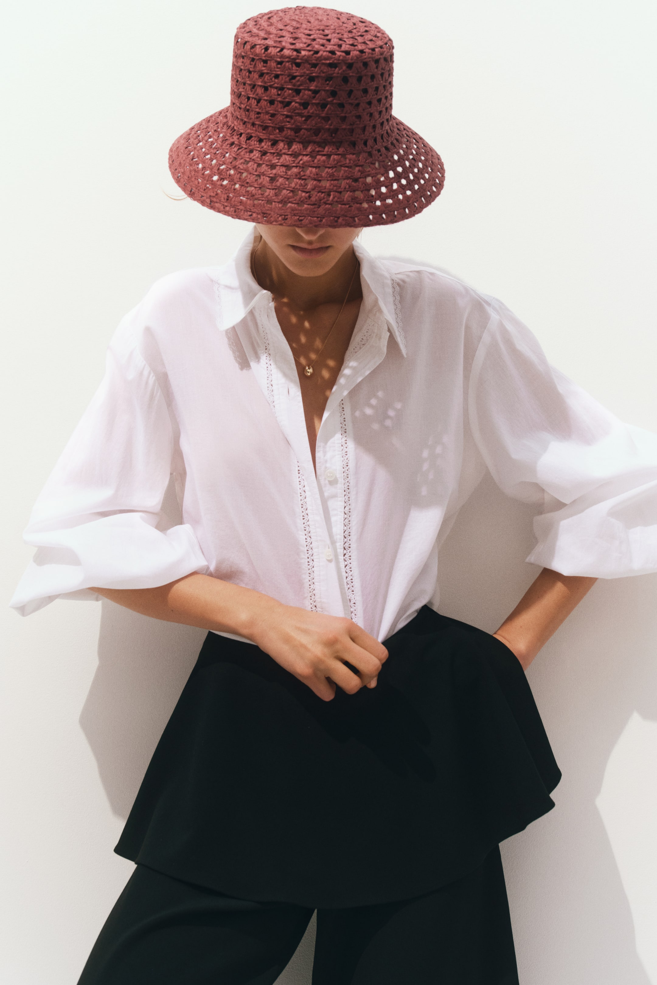 Visualizza immagine più grande: Il prodotto presenta un cappello a secchiello a trama aperta di colore rosso scuro, una camicia bianca oversize con cannoncino rifinito in pizzo e maniche a sbuffo, e pantaloni neri a gamba larga con dettaglio a peplo stratificato, tutti presentati da un'angolazione frontale.