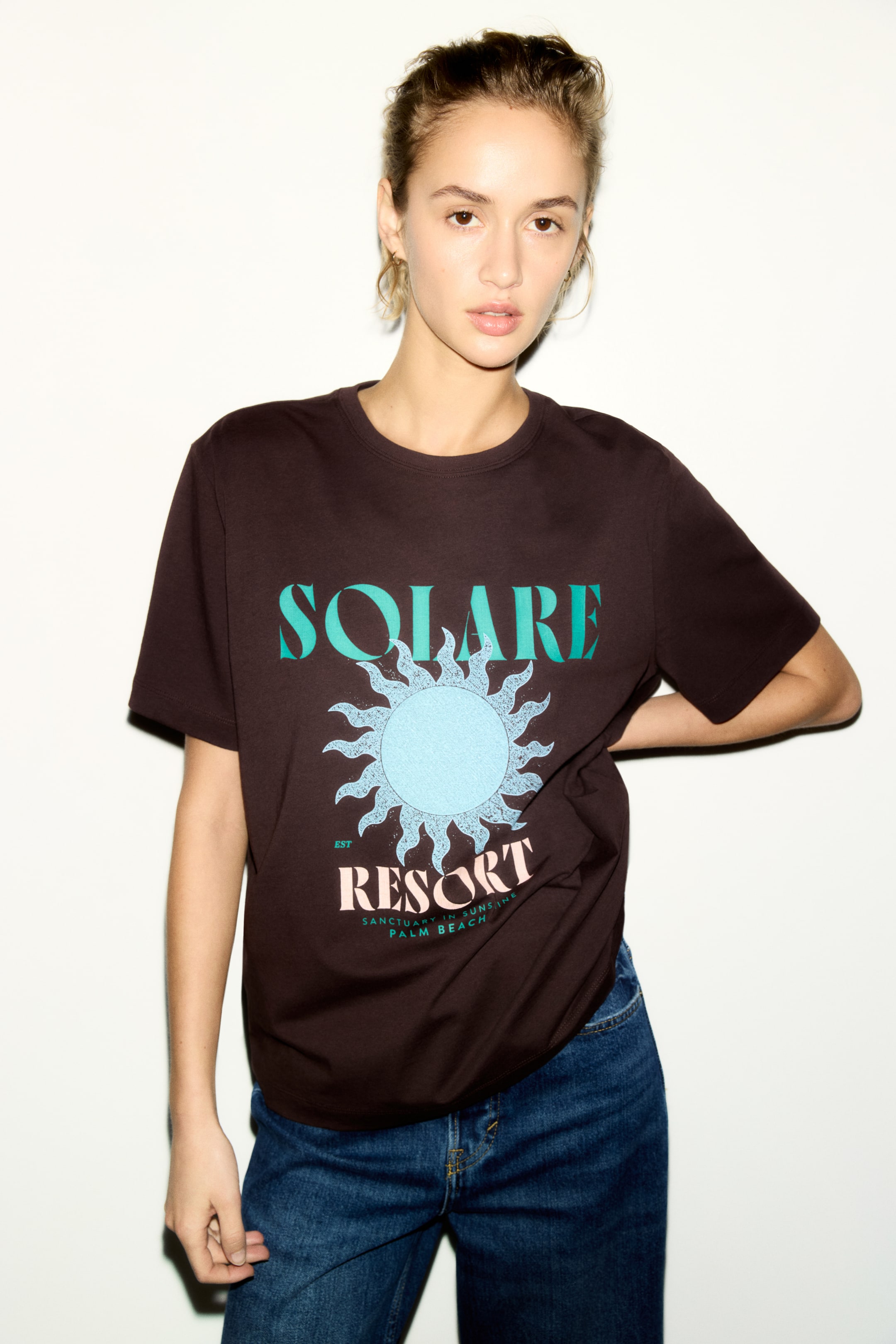 View larger image: The dark brown short-sleeve t-shirt showcases a prominent graphic on the front, facing outward. The design includes "SOLARE" in teal, a light blue sun motif with wavy rays, and "RESORT" in pink, accompanied by smaller teal text below.