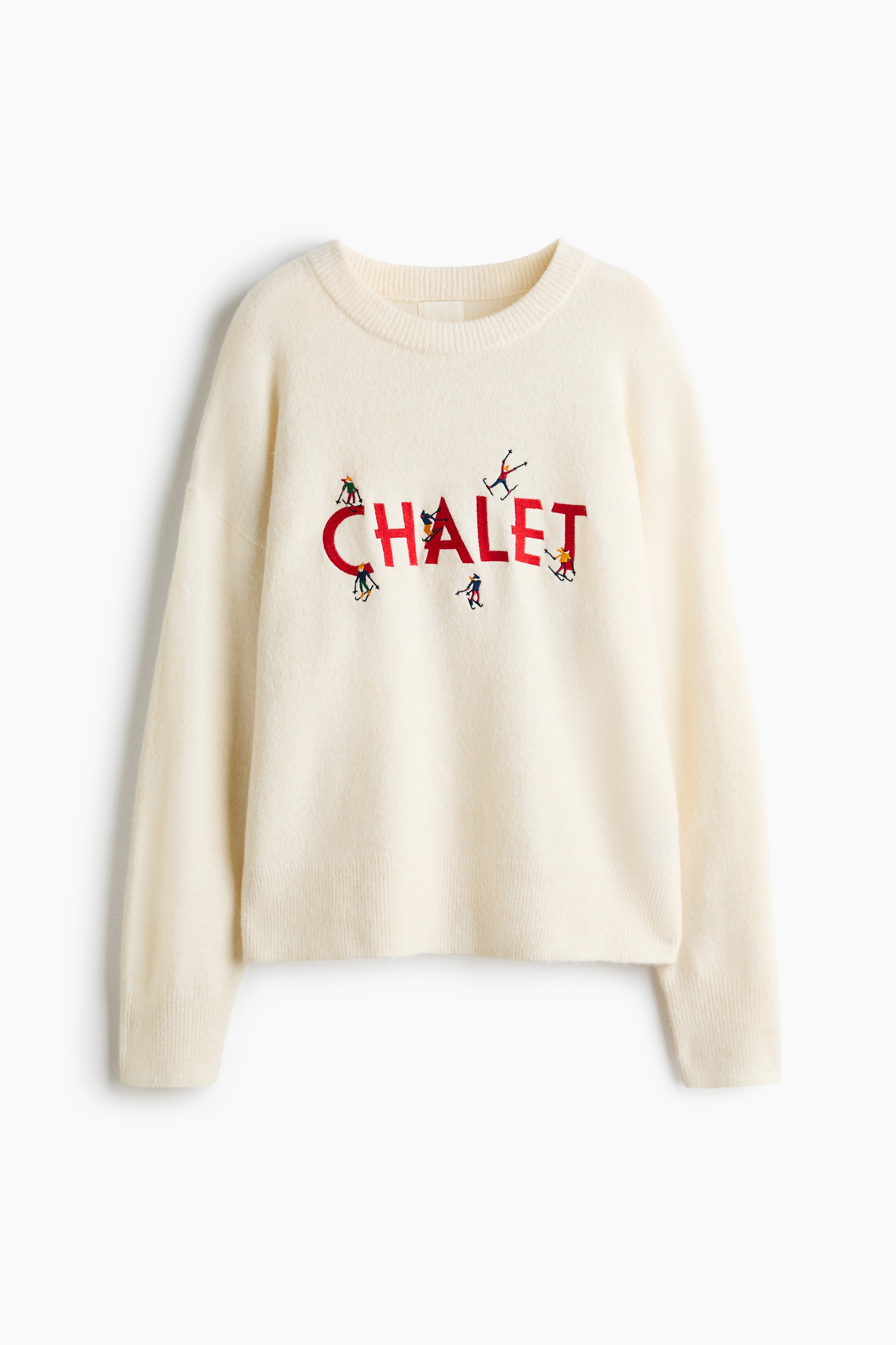 La modelo lleva un suéter tejido color crema de corte oversized, con cuello redondo acanalado, hombros caídos y un gráfico bordado en rojo en el pecho. La modelo sostiene los extremos drapeados de una prenda tejida a juego, anudada a la cintura, combinada con jeans azules y un cinturón negro.