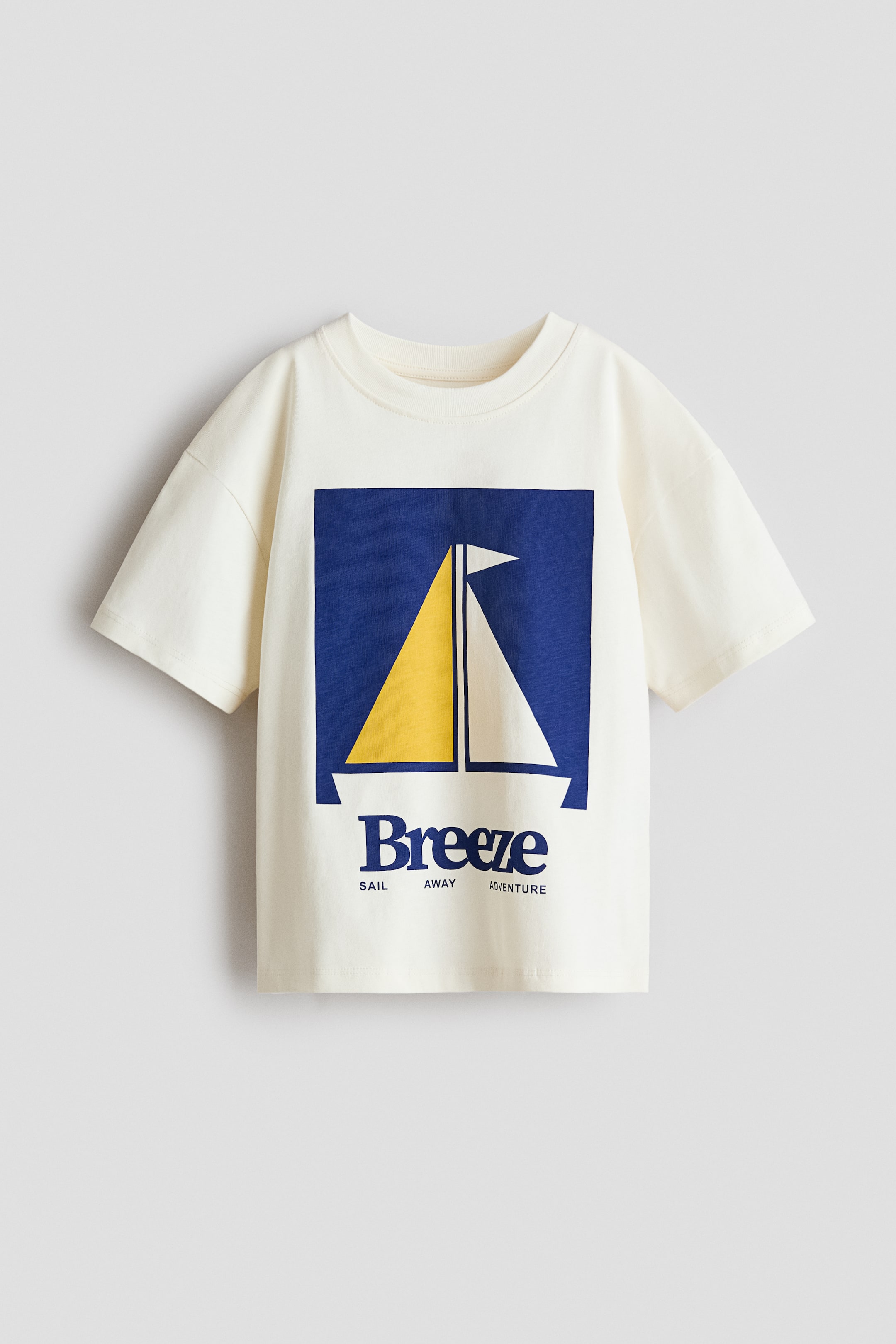 Visualizza l'immagine ingrandita: Una t-shirt a vestibilità ampia in morbido jersey di cotone bianco sporco è presentata dal davanti, caratterizzata da uno scollo rotondo con bordo a coste, spalle scese e maniche corte. Un motivo stampato sul davanti raffigura una barca a vela stilizzata con vele gialle e bianche all'interno di un rettangolo blu, accompagnata dalle parole "Breeze", "SAIL AWAY" e "ADVENTURE."
