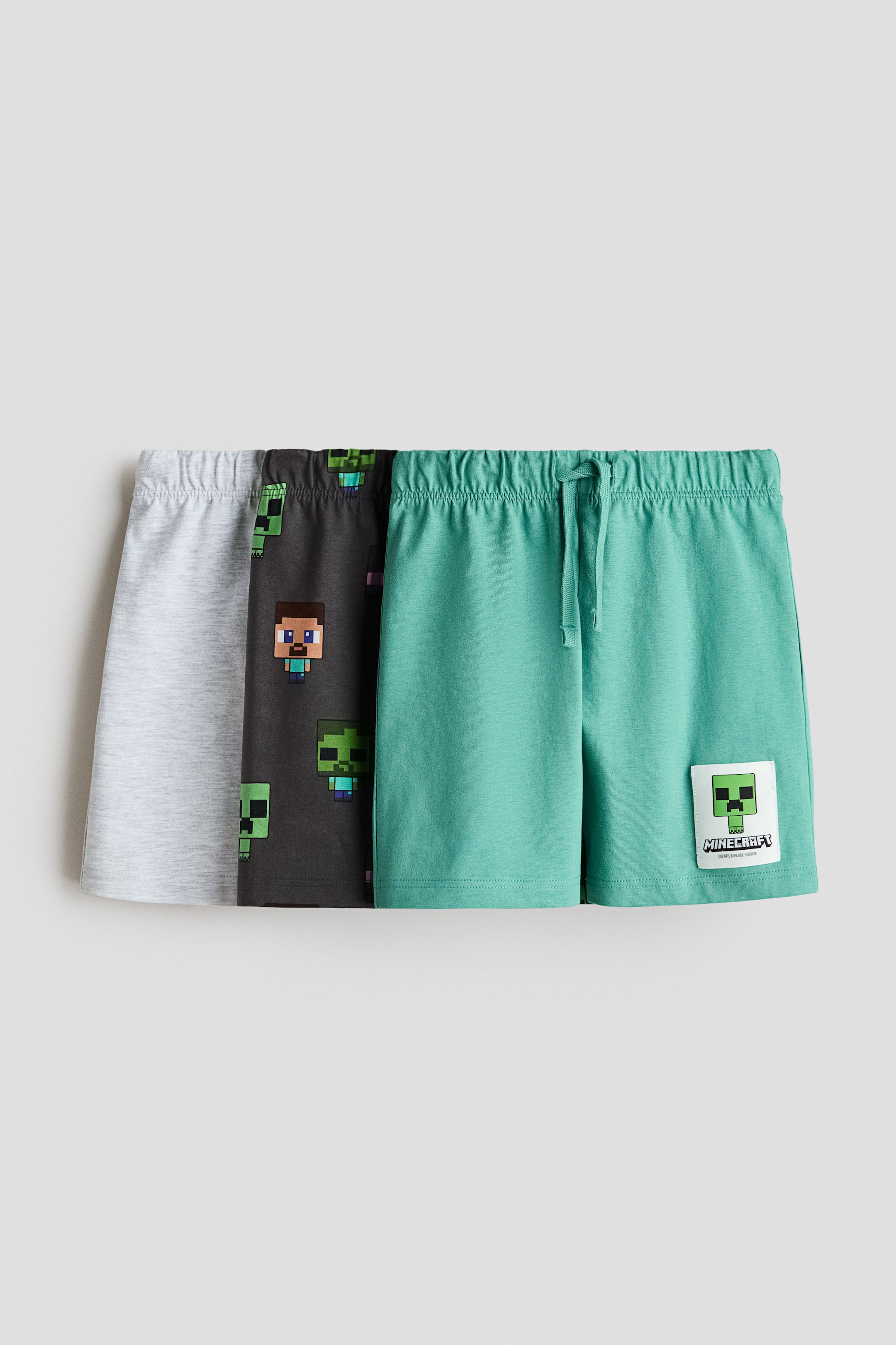View larger image: Three pairs of children's shorts are showcased from the front, layered together. The left short is in a light grey marl, the middle dark grey short features a repeating Minecraft character print, and the right teal short displays a Minecraft Creeper patch on the leg with an elasticated drawstring waistband.