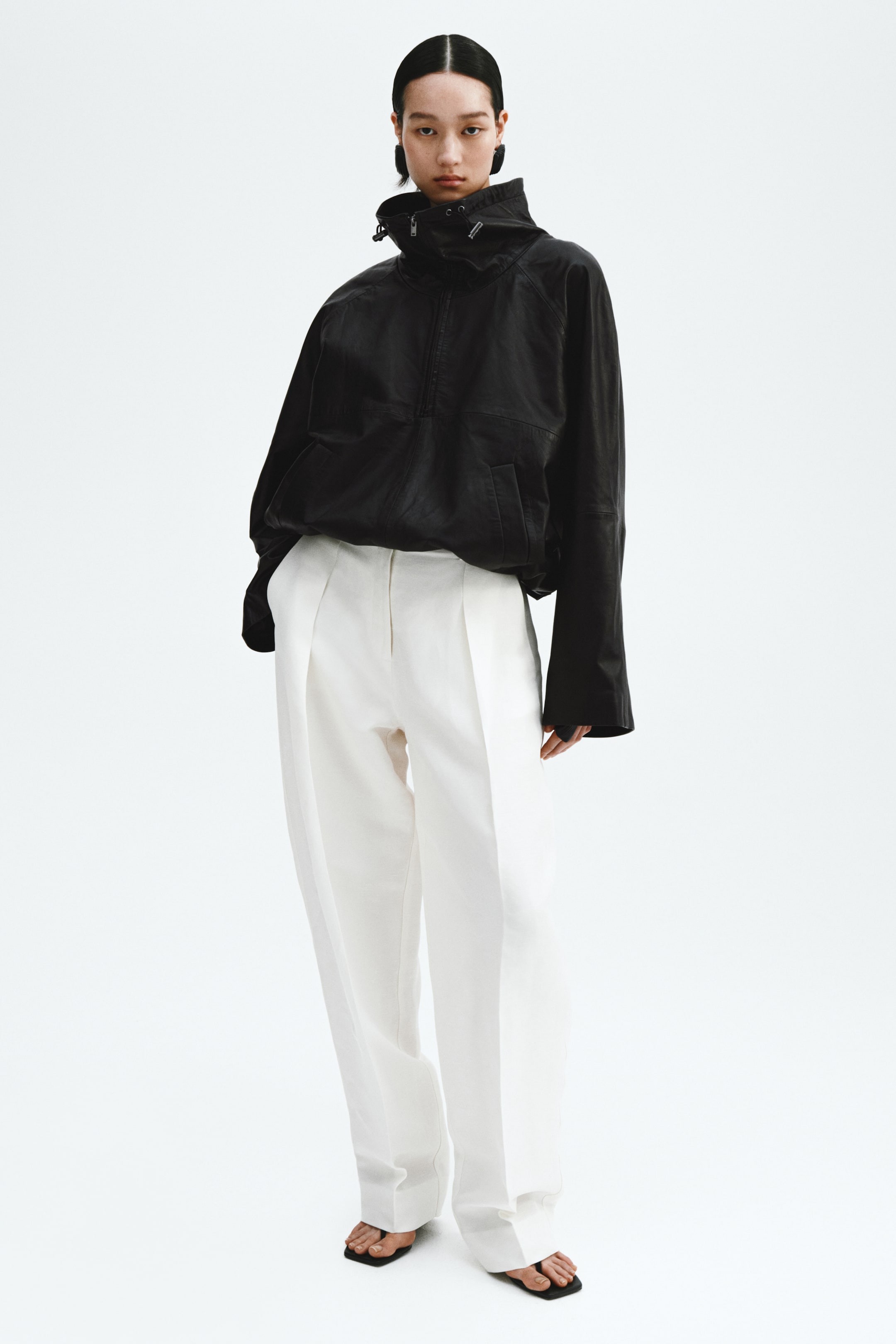 Se større billede: En sort pullover anorak med høj snørekrave, halv lynlås og brede ærmer bæres over hvide plisserede bukser med høj talje, designet med vidde ben.