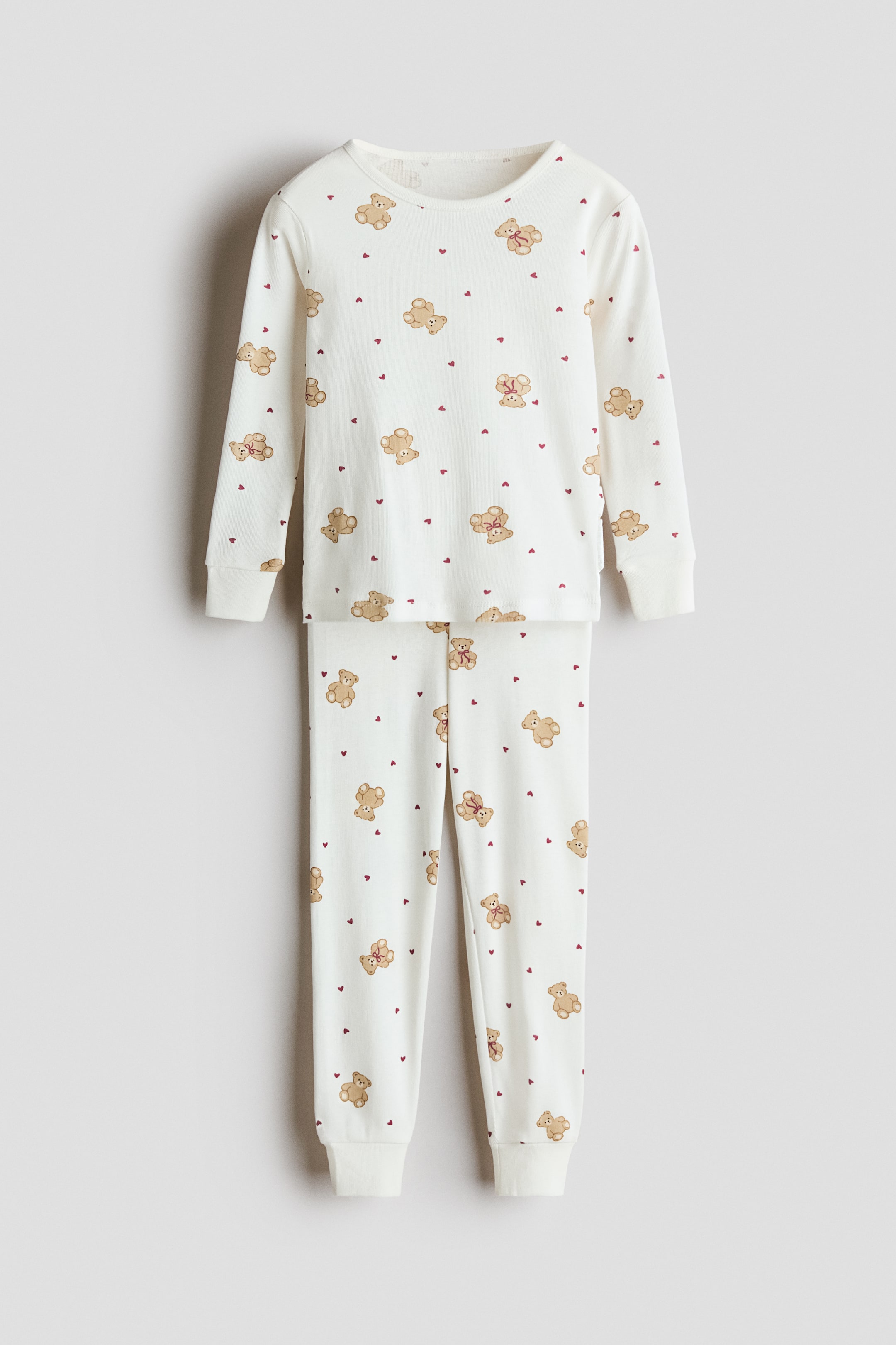 View larger image: A pair of pajamas is shown from the front, featuring an off-white top with a round neckline, long sleeves, and ribbed cuffs, along with matching bottoms with an elastic waistband and ribbed hems. Both pieces are covered in a pattern of brown teddy bears and small red hearts.