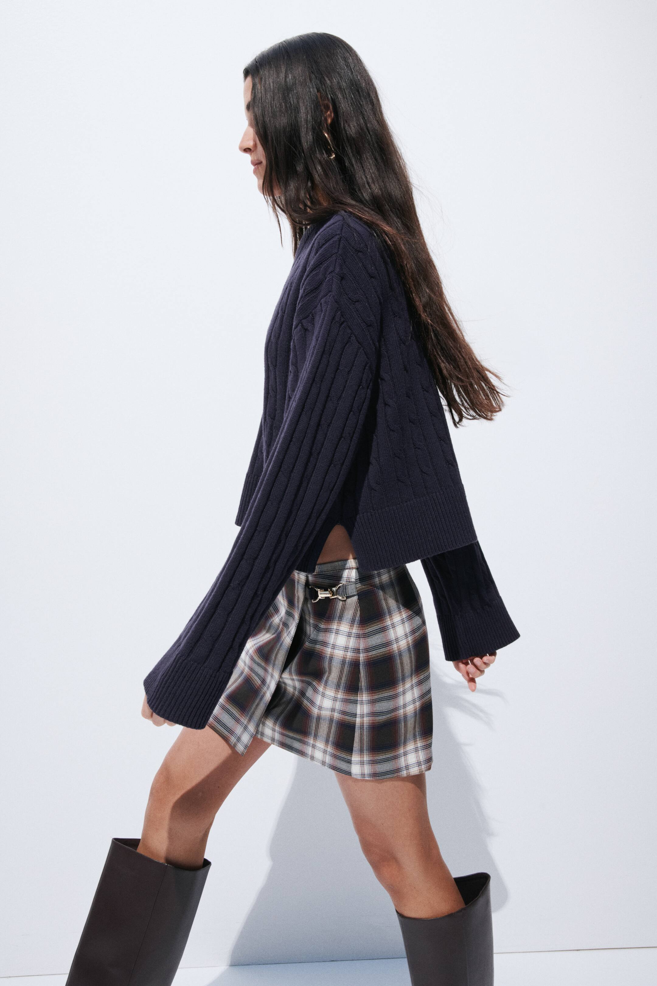 View larger image: The navy blue cable knit jumper showcases a loose, cropped fit with side slits, a cable knit pattern, and long, wide sleeves with ribbed cuffs. It is presented from a side angle.