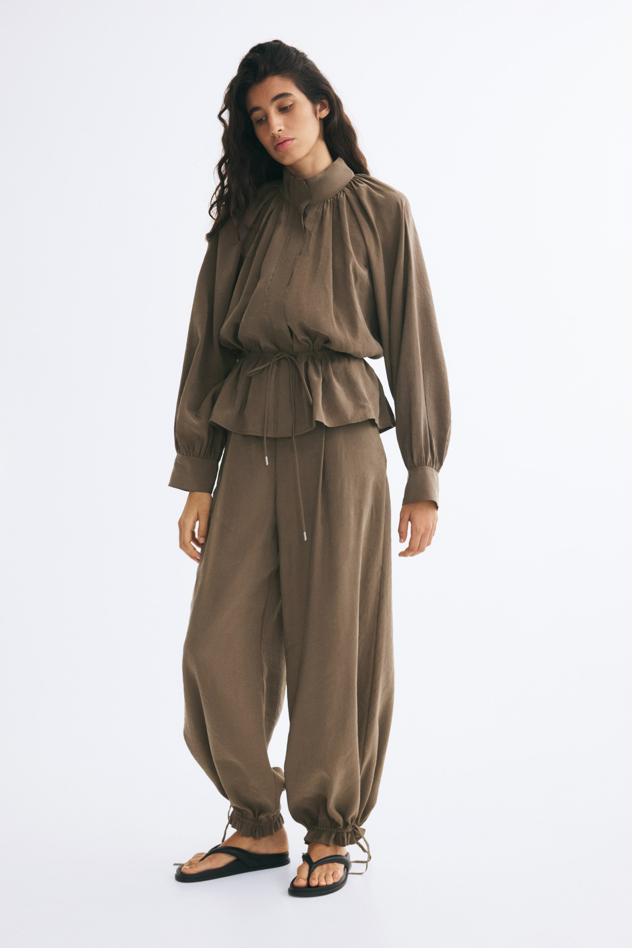 View larger image: Loose-fit brown pants in a lightly crinkled fabric are worn by the model, featuring balloon legs with narrow, adjustable ties gathered at the hems. The pants are paired with a matching long-sleeve, high-collar top and black flat sandals.