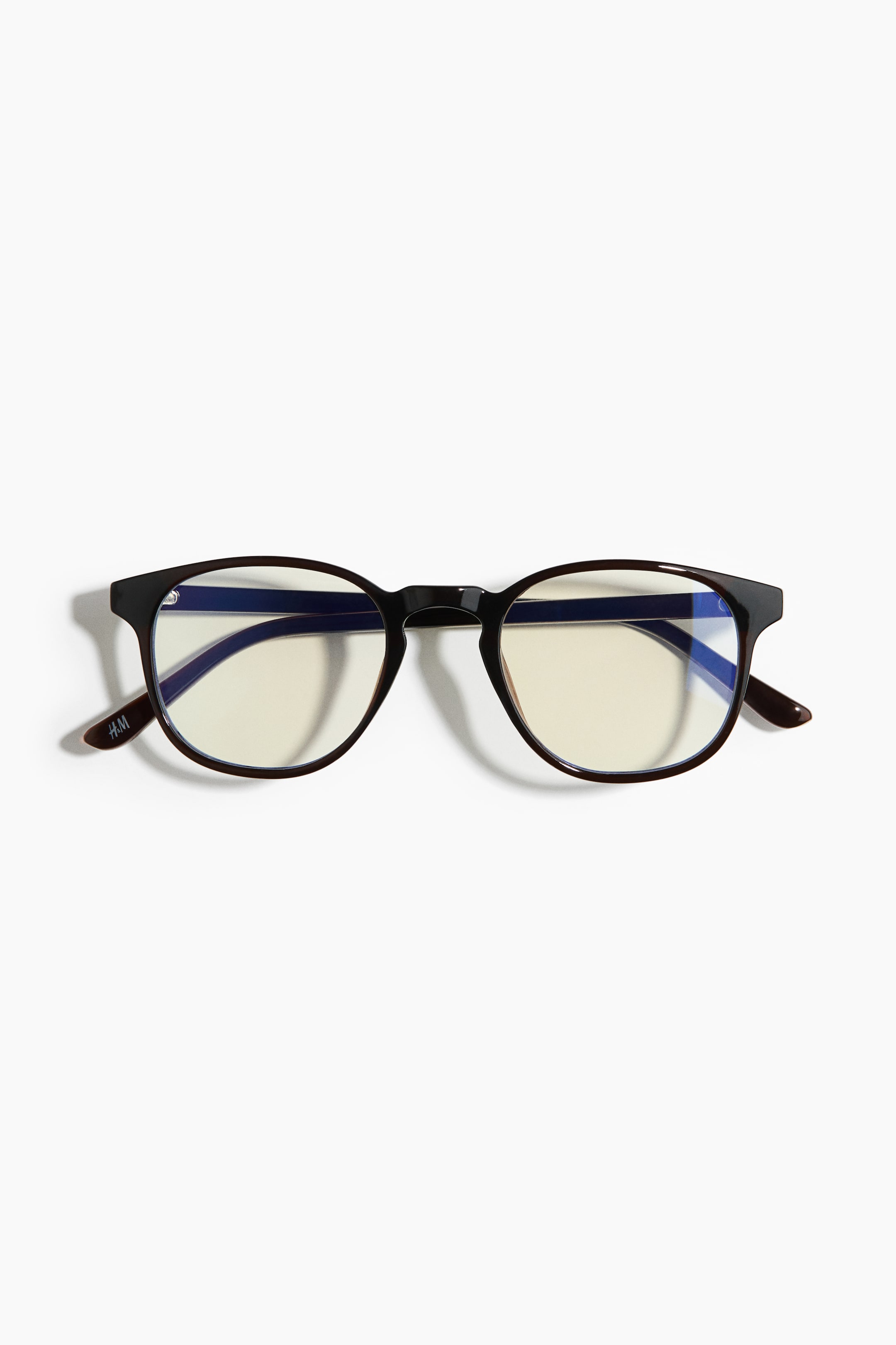 Agrandir l'image: Les lunettes sont posées à plat, orientées vers l'avant, avec des montures pleines de couleur brun foncé et des verres ronds affichant une légère teinte jaunâtre et des reflets bleus. Le logo H&M est visible sur la branche gauche.