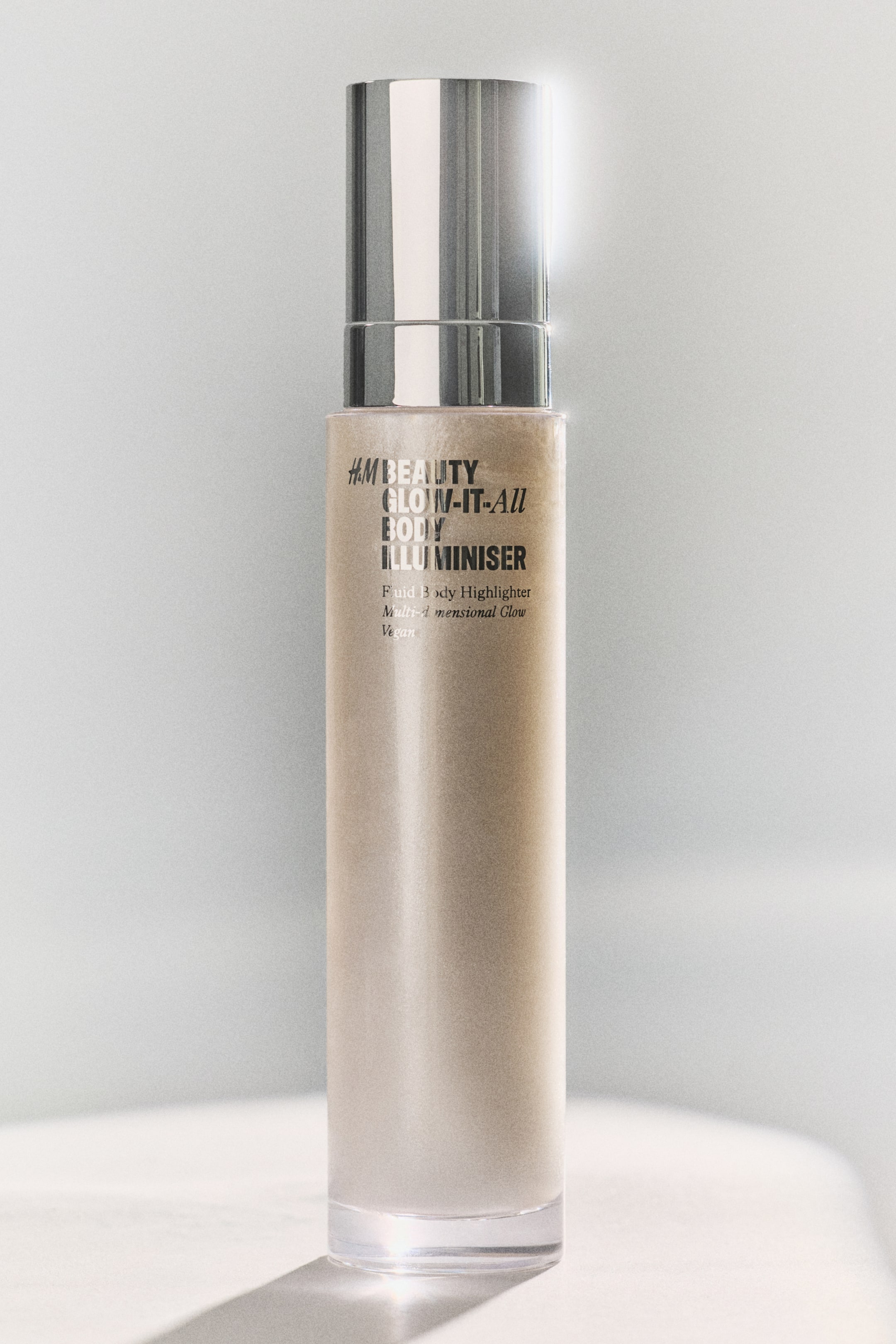 View larger image: An H&M Beauty Glow-It-All Body Illuminiser is standing upright, featuring a tall, cylindrical clear bottle filled with a shimmering, light gold liquid and topped with a reflective silver cap. The label on the bottle is displaying "H&M BEAUTY GLOW-IT-ALL BODY ILLUMINISER," "Fluid Body Highlighter," "Multi-dimensional Glow," and "Vegan" in black text.