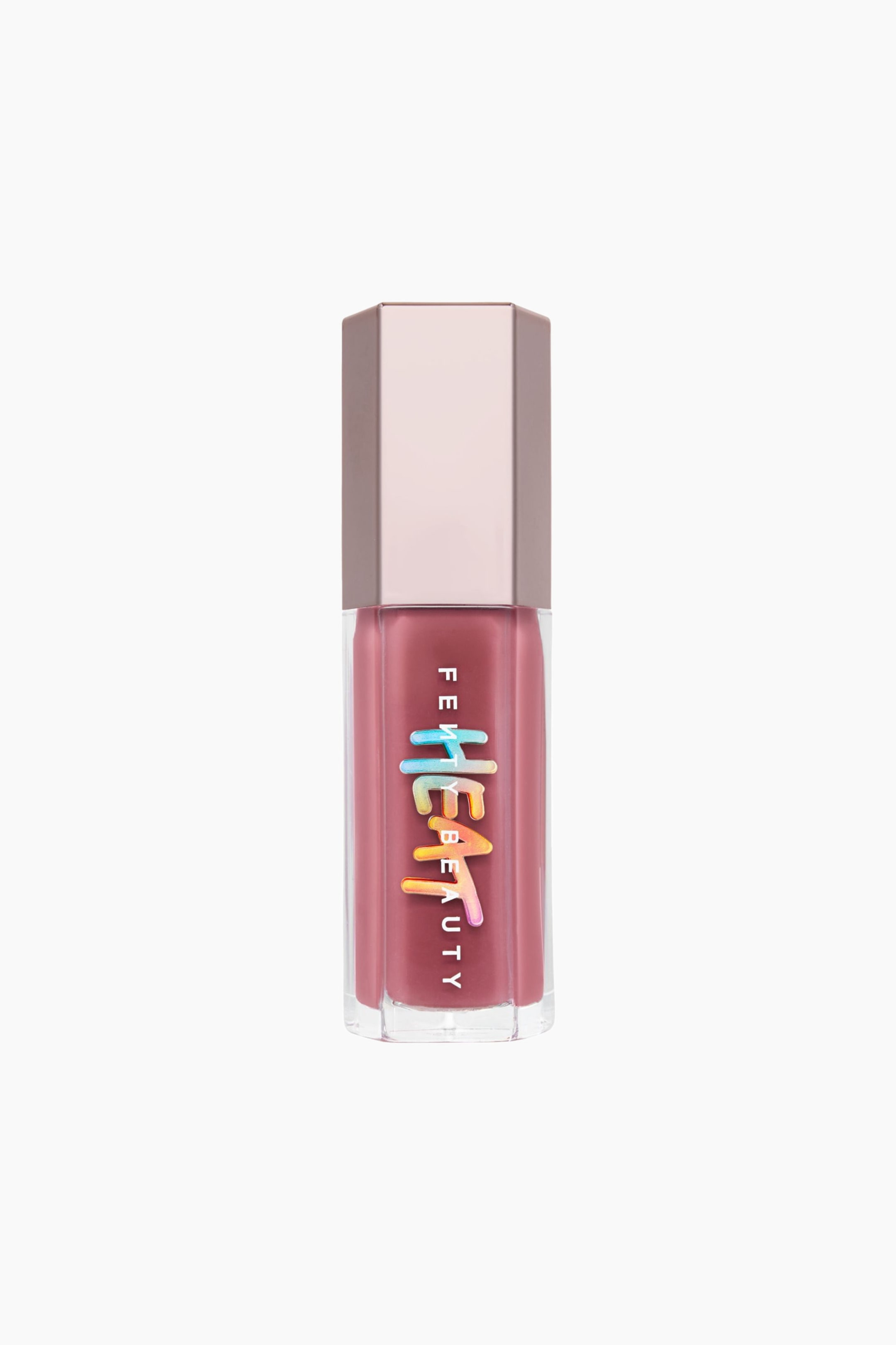 Visa större bild: Gloss Bomb Heat Universal Lip Luminizer + Plumper står upprätt och har en genomskinlig sexkantig tub fylld med ett dammrosa gloss, toppad med ett metalliskt roséguldfärgat sexkantigt lock. Framsidan av behållaren visar den vertikala vita "FENTY BEAUTY"-logotypen överlagrad med en stiliserad, droppande "HEAT"-grafik i en flerfärgad gradient.
