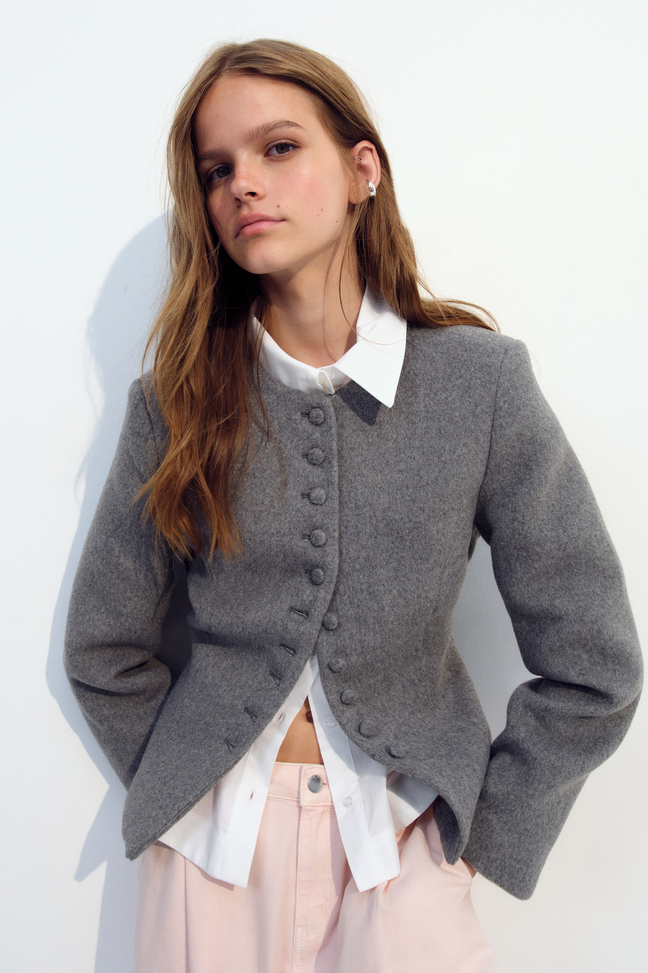Ampliar la imagen: La modelo lleva puesta una chaqueta gris texturizada con cuello redondo sin solapas y una hilera de botones forrados en tela. La chaqueta se lleva sobre una camisa blanca con cuello y se combina con pantalones rosa claro.