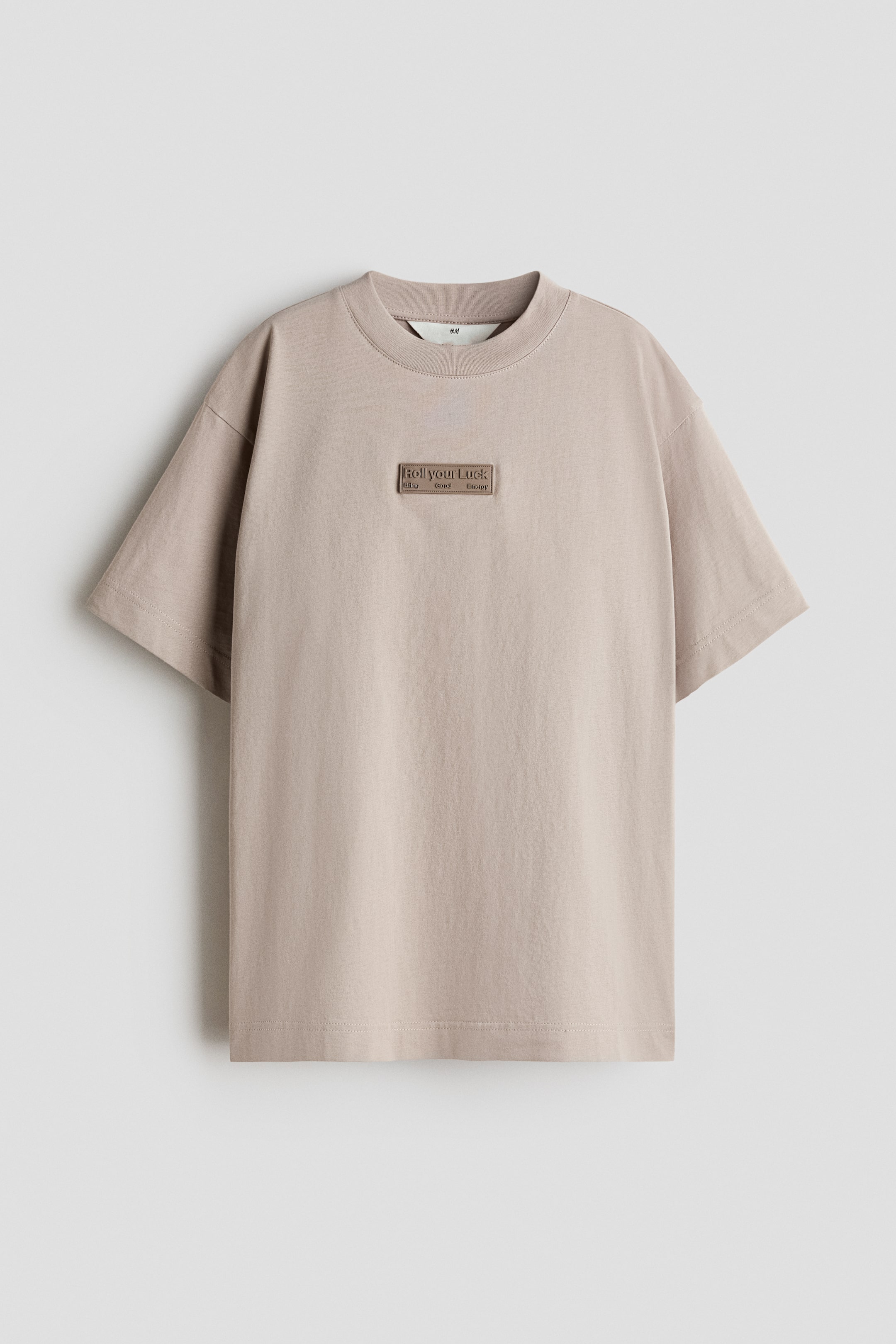Agrandir l'image: Un t-shirt beige clair, coupe ample, en jersey de coton doux, se présente de face, avec un col rond bordé de côtes et des épaules légèrement tombantes. Un motif embossé rectangulaire est placé au centre à l'avant, affichant le texte "Roll your Luck" au-dessus de "living good energy".