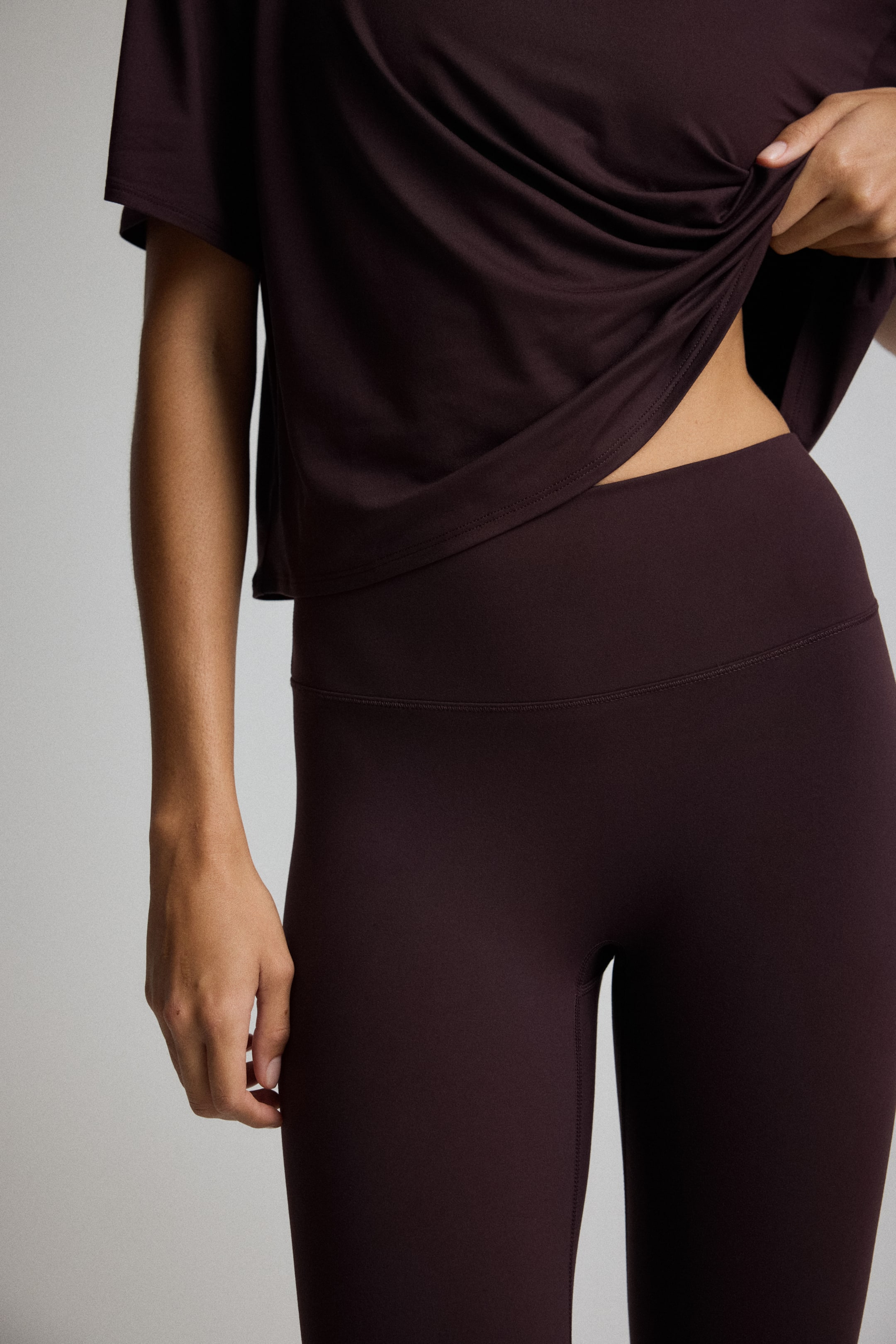 View larger image: Dark-colored, high-waisted leggings are fitting snugly, with their wide, smooth waistband visible from a slight side angle. A matching short-sleeved top is partially covering the leggings, and its hem is being lifted by a hand, revealing the top portion of the leggings.
