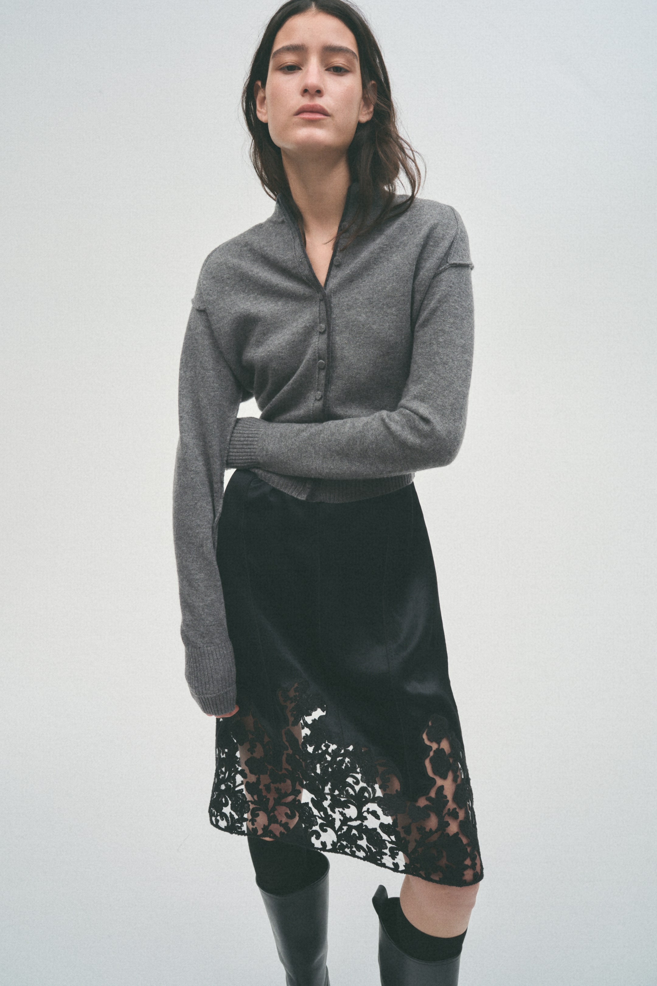 View larger image: A grey V-neck cardigan with small buttons is worn by the model, layered over a dark top. The model is also wearing a black skirt featuring a satin-like texture and a wide floral lace trim at the hem.