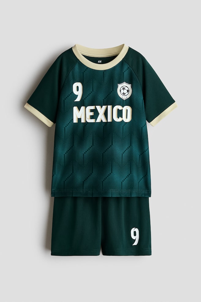 A printed soccer set in soft mesh fabric is shown facing front, featuring a dark green short-sleeve top and matching shorts. The top is displaying a textured dot pattern, a cream-colored crew neck and sleeve cuffs, a soccer crest, and the text "9 MEXICO" on the chest, while the shorts are featuring a printed number "9" on the left leg. - 1