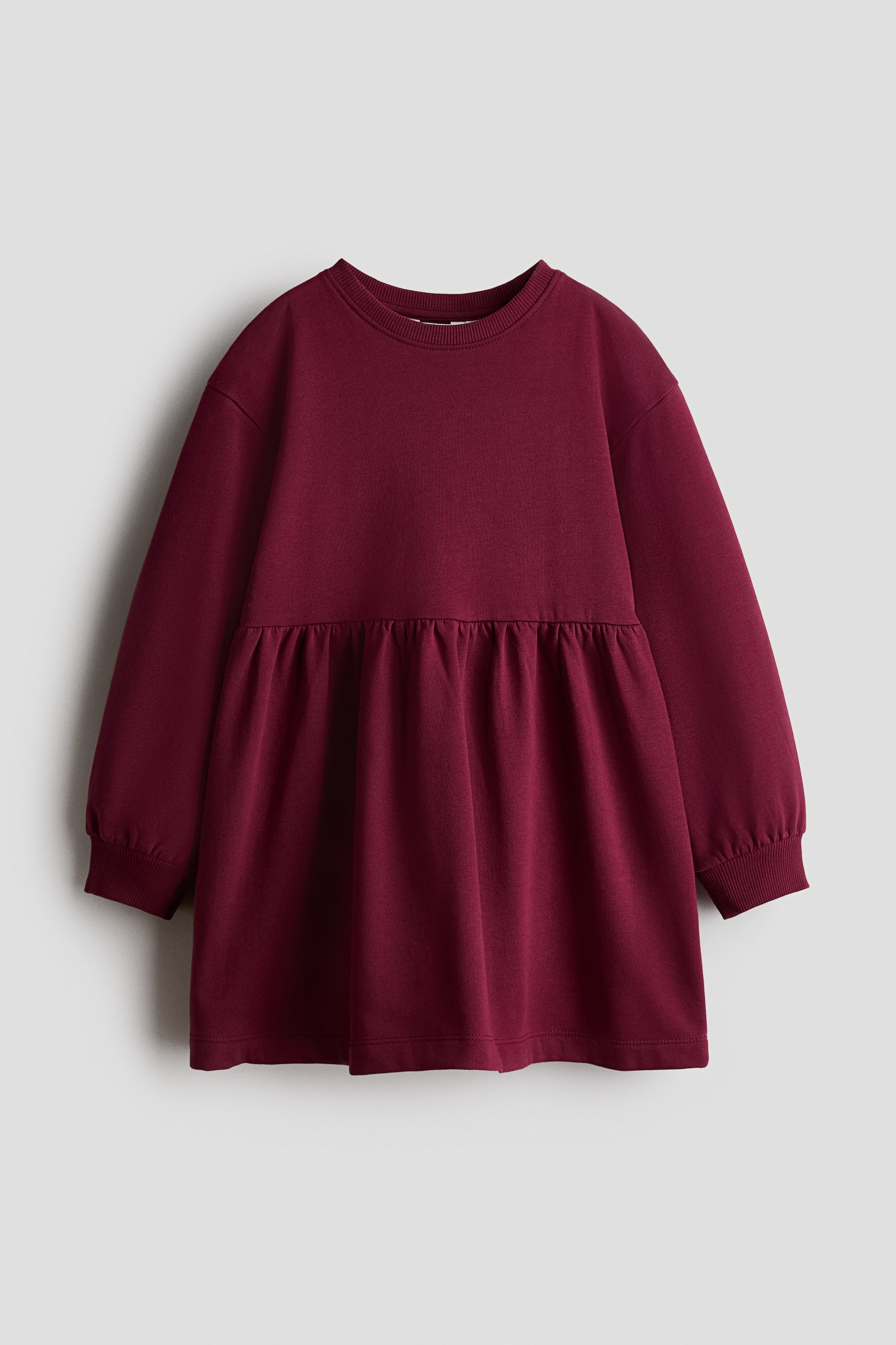 Näytä suurempi kuva: Burgundinpunainen, väljä fleece-mekko on kuvattuna edestä päin. Siinä on pyöreä, resoreilla viimeistelty pääntie, laskettu olkalinja ja pitkät hihat resorimanseteilla. Poimutettu sauma korostaa korkeaa vyötäröä, josta helma levenee pehmeästi.