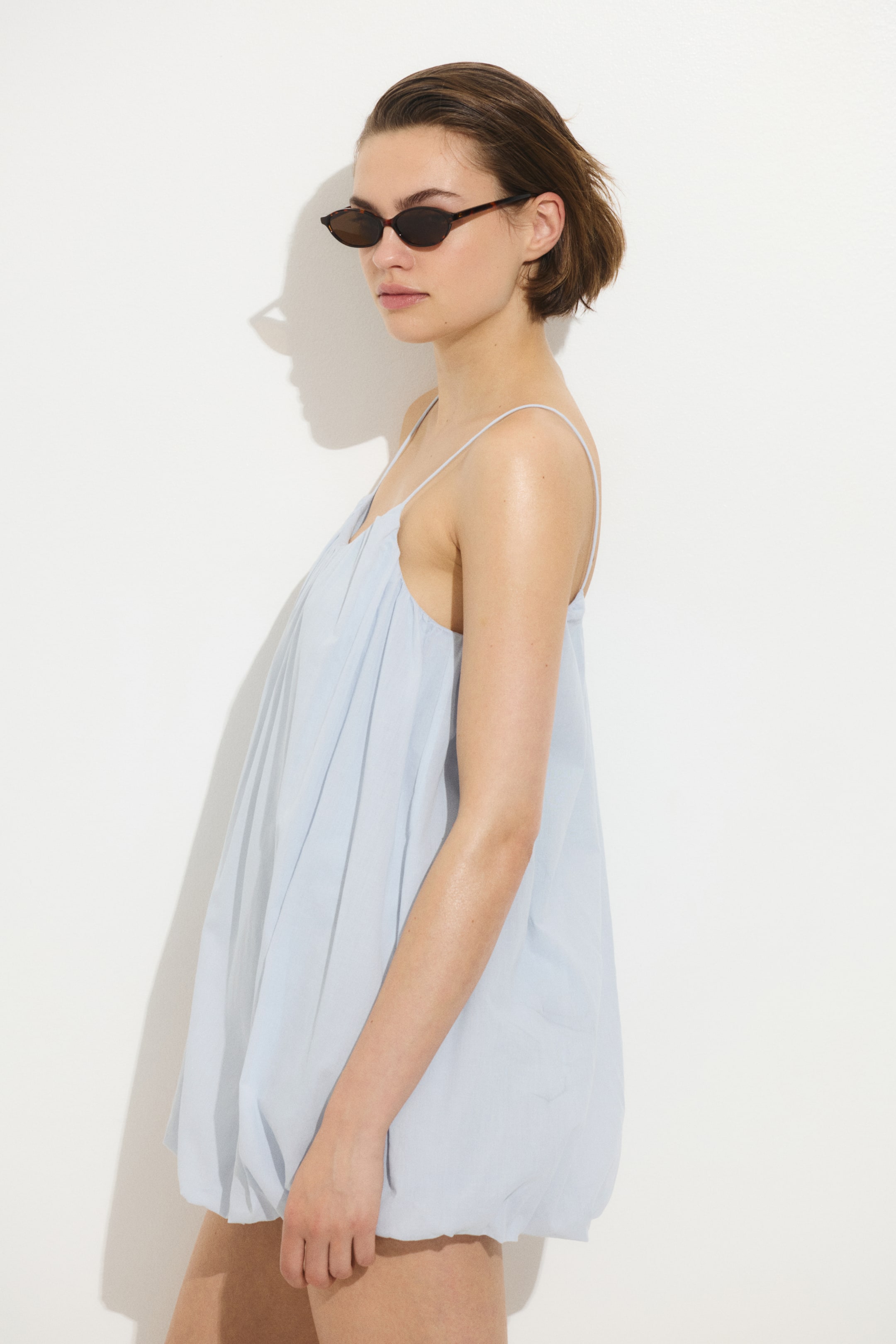 拡大画像を表示: モデルが着用しているのは、スパゲッティショルダーストラップ、ギャザースクープネックライン、そしてふんわりとしたシルエットを作り出すバブルヘムが特徴のライトブルーのミニドレスです。