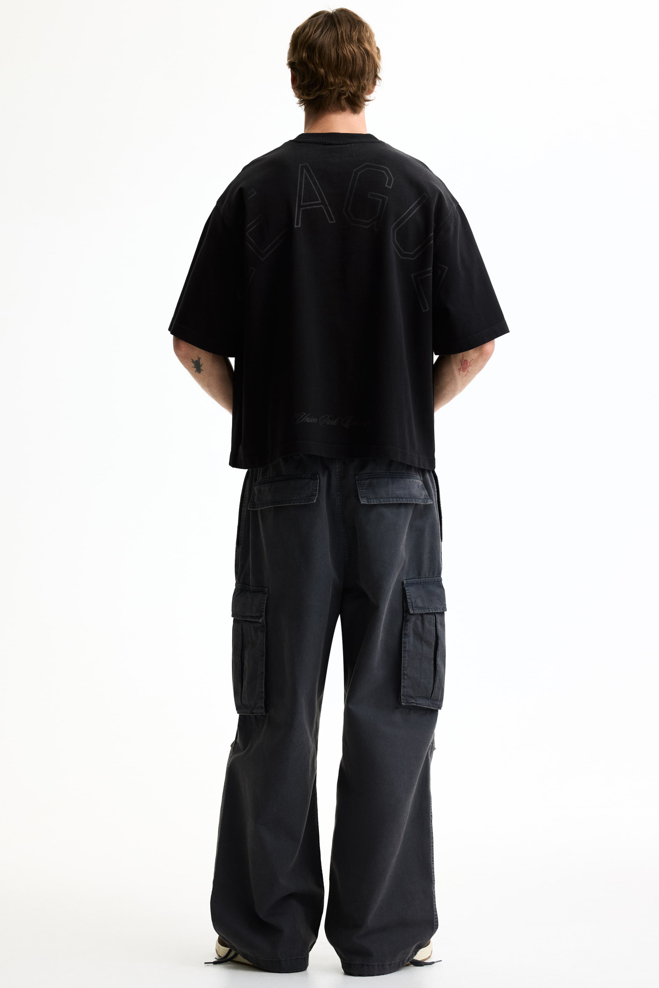 Agrandir l'image: Un t-shirt noir oversize est présenté de dos, avec le texte discret "LEAGUE" sur le haut du dos et une petite inscription au-dessus de son ourlet raccourci. Il est porté avec un pantalon cargo noir ample, doté de plusieurs poches à rabat sur les côtés.