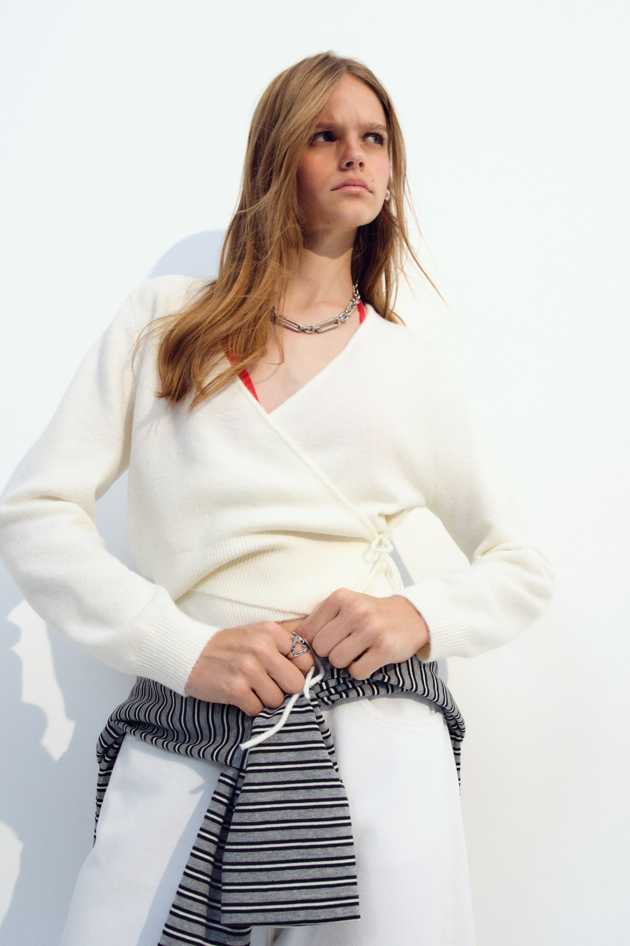 Ampliar la imagen: La modelo lleva un cárdigan corto cruzado color crema, de tejido suave, con escote en V y puños acanalados. El cárdigan se combina con una blusa roja y pantalones blancos, mientras la modelo sostiene las mangas de un suéter a rayas amarrado a su cintura.