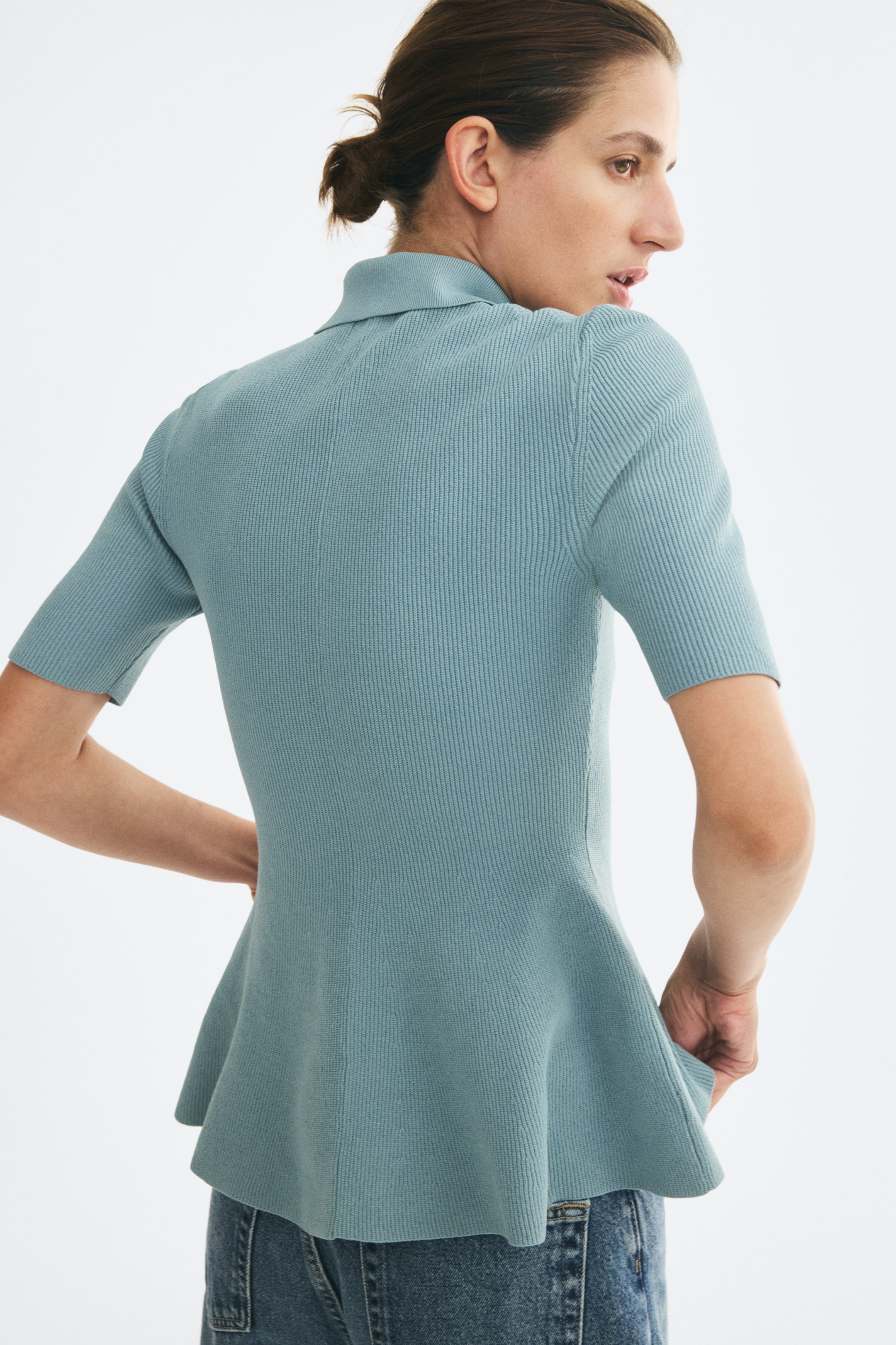 Xem ảnh lớn hơn: Người mẫu đang mặc một chiếc áo polo dệt gân màu xanh lam - xanh lục nhạt, có tay ngắn, cổ bẻ và gấu peplum xòe. Áo được phối cùng quần jeans xanh, trong tư thế người mẫu đứng quay lưng nghiêng một phần.