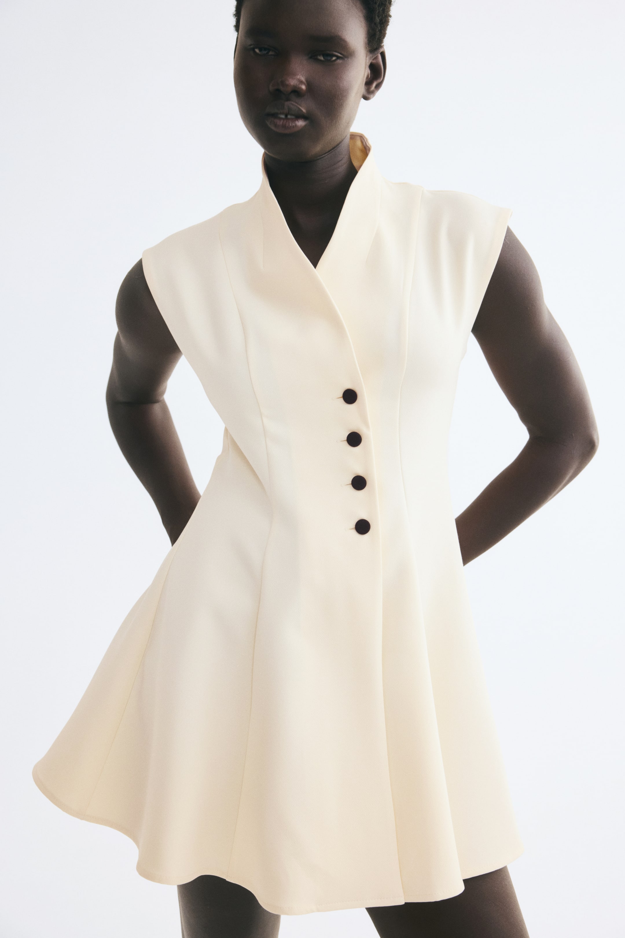 Größeres Bild ansehen: Ein cremefarbenes, kurzes Wickelkleid wird von dem Model getragen und verfügt über einen strukturierten Stehkragen, einen V-Ausschnitt und einen vorderen Verschluss mit vier dunklen Knöpfen. Das ärmellose Kleid hat vertikale formgebende Nähte an der Vorderseite, die in einen ausgestellten Rock übergehen.