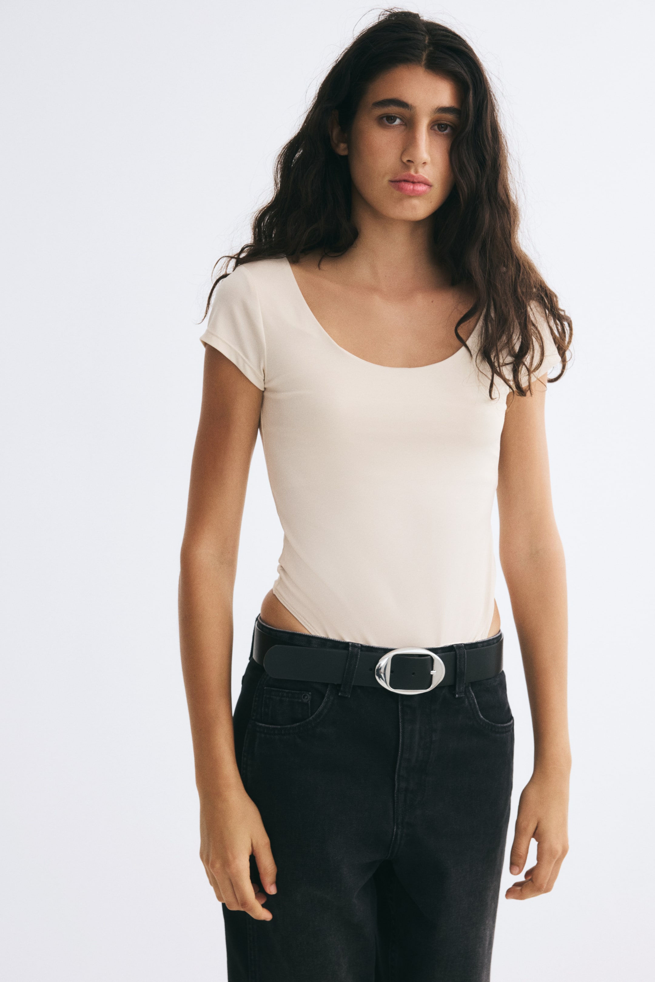 View larger image: A light beige, short-sleeve jersey bodysuit with a scoop neckline is worn by the model. The bodysuit is styled tucked into black jeans with a black belt, featuring a high-cut hip detail above the waistband.