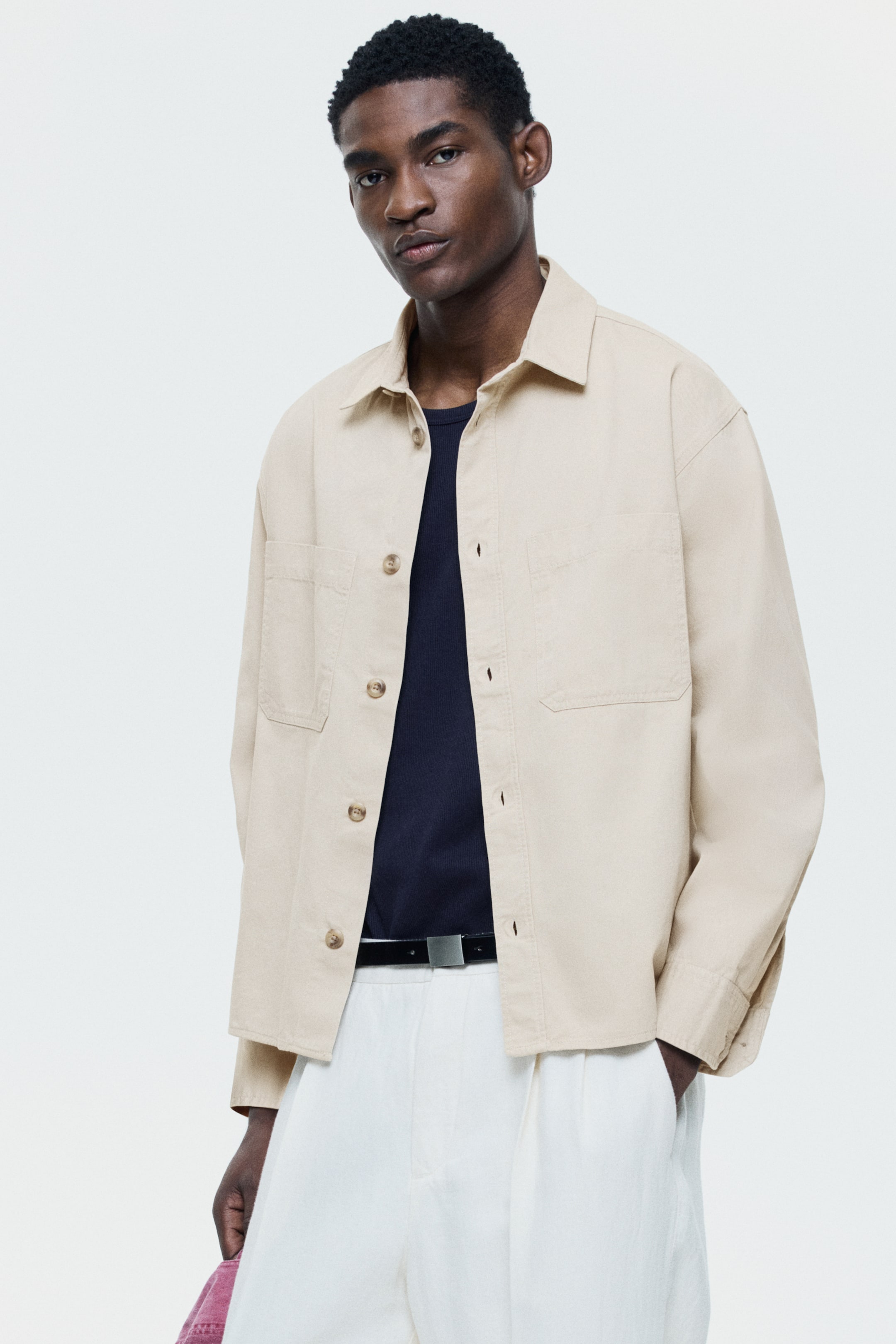 View larger image: A beige overshirt is showcased from a slight front-right angle, featuring a pointed collar and two large patch pockets on the chest. It is designed with a front button closure, highlighting light brown buttons, and is layered over a dark navy t-shirt.