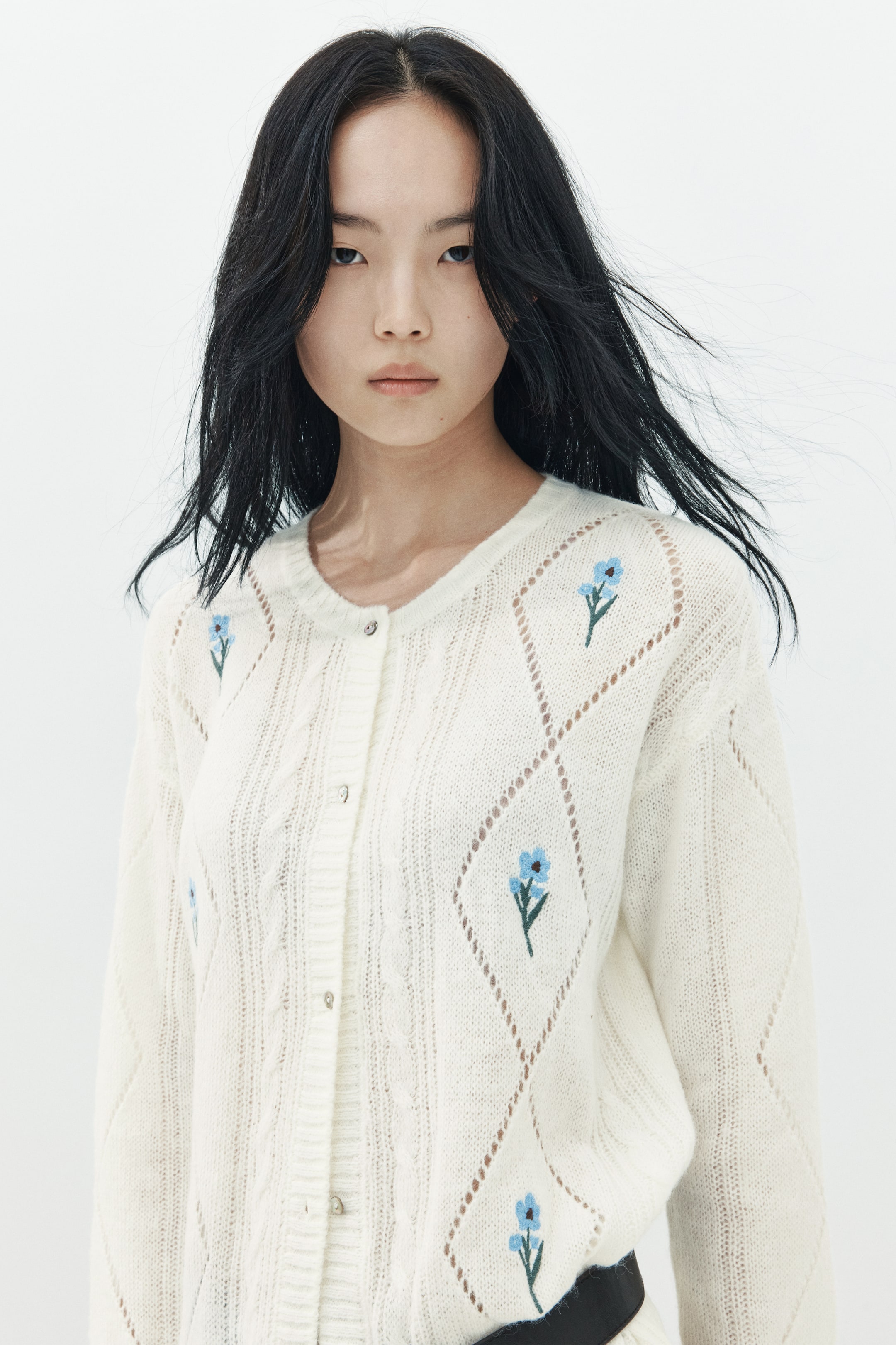 View larger image: The model is wearing an off-white knit cardigan featuring embroidered blue flowers within diamond-shaped pointelle patterns. The cardigan is styled with a button-front closure, crew neck, and subtle cable-knit textures.