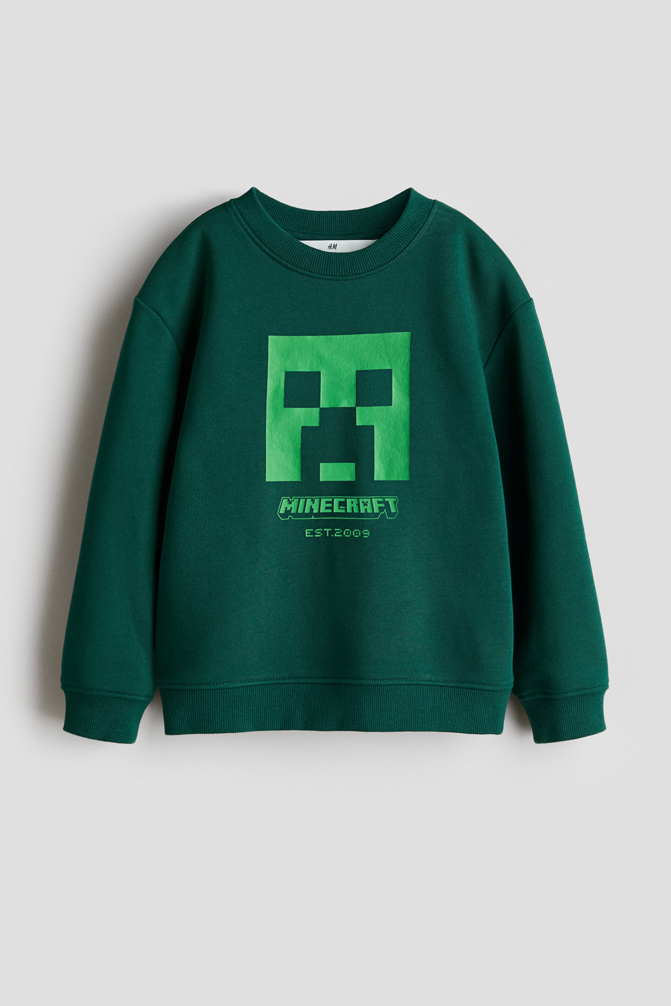 Ver imagen más grande: Una sudadera verde oscuro está orientada hacia el FRENTE, con un gran estampado pixelado de cara de Creeper en verde claro en el pecho y el texto "MINECRAFT EST.2009" debajo. Está diseñada con un cuello redondo con ribete, hombros caídos, mangas largas y detalles de canalé en los puños y el dobladillo.
