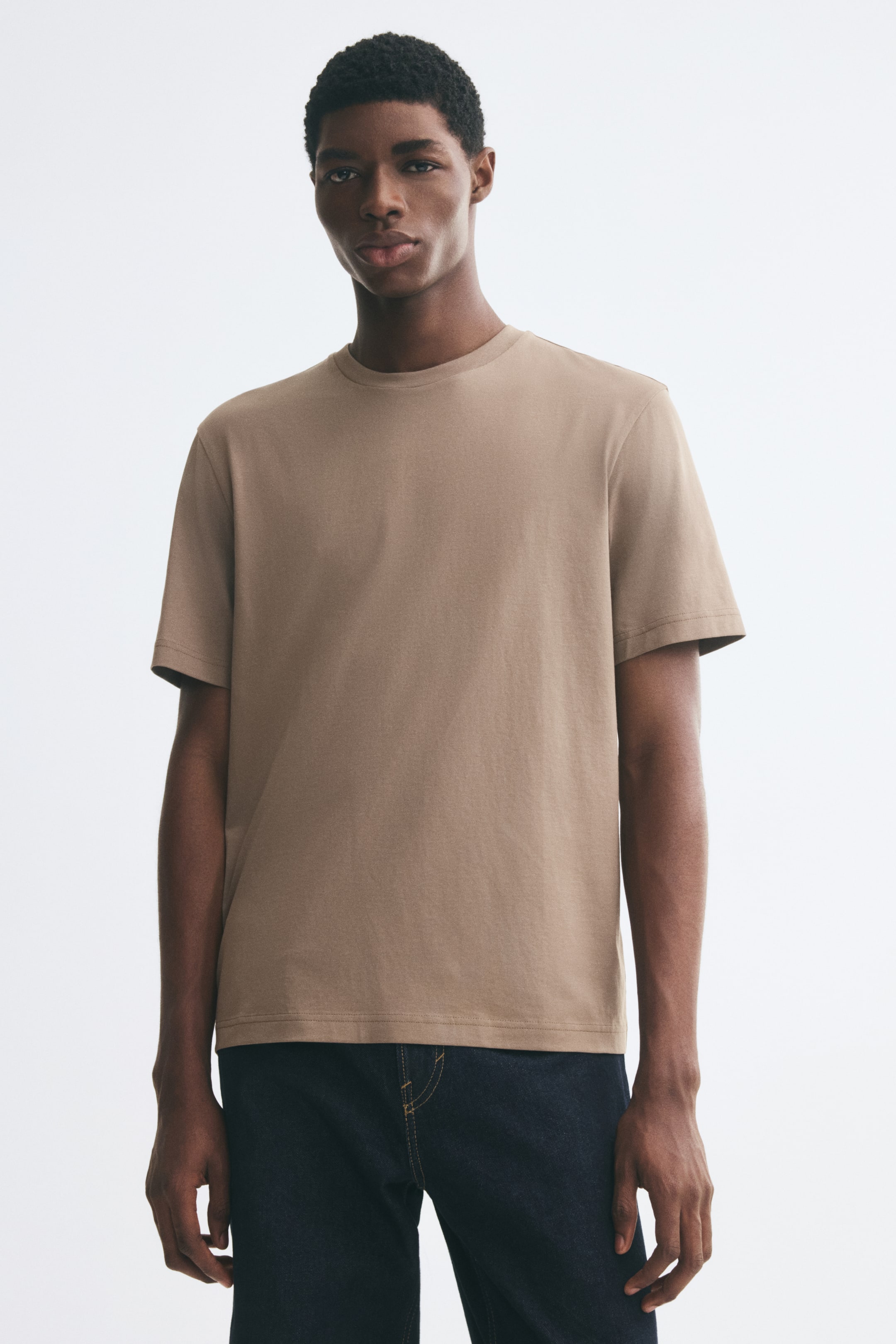 Größeres Bild anzeigen: Das Model trägt ein beiges T-Shirt mit kurzen Ärmeln und Rundhalsausschnitt. Das unifarbene T-Shirt hat eine Regular Fit-Passform und wird mit dunkel gewaschenen Jeans kombiniert.