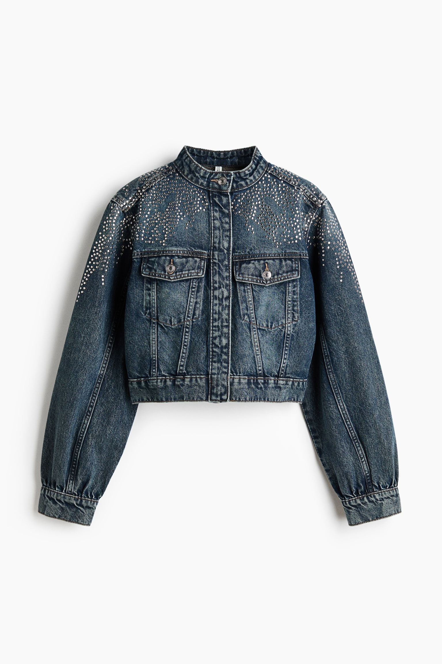 A cropped blue denim jacket is shown from the front, featuring a stand collar with a single visible button and a concealed front fastening. Small silver studs embellish the shoulders and upper chest, while buttoned flap pockets detail the front.