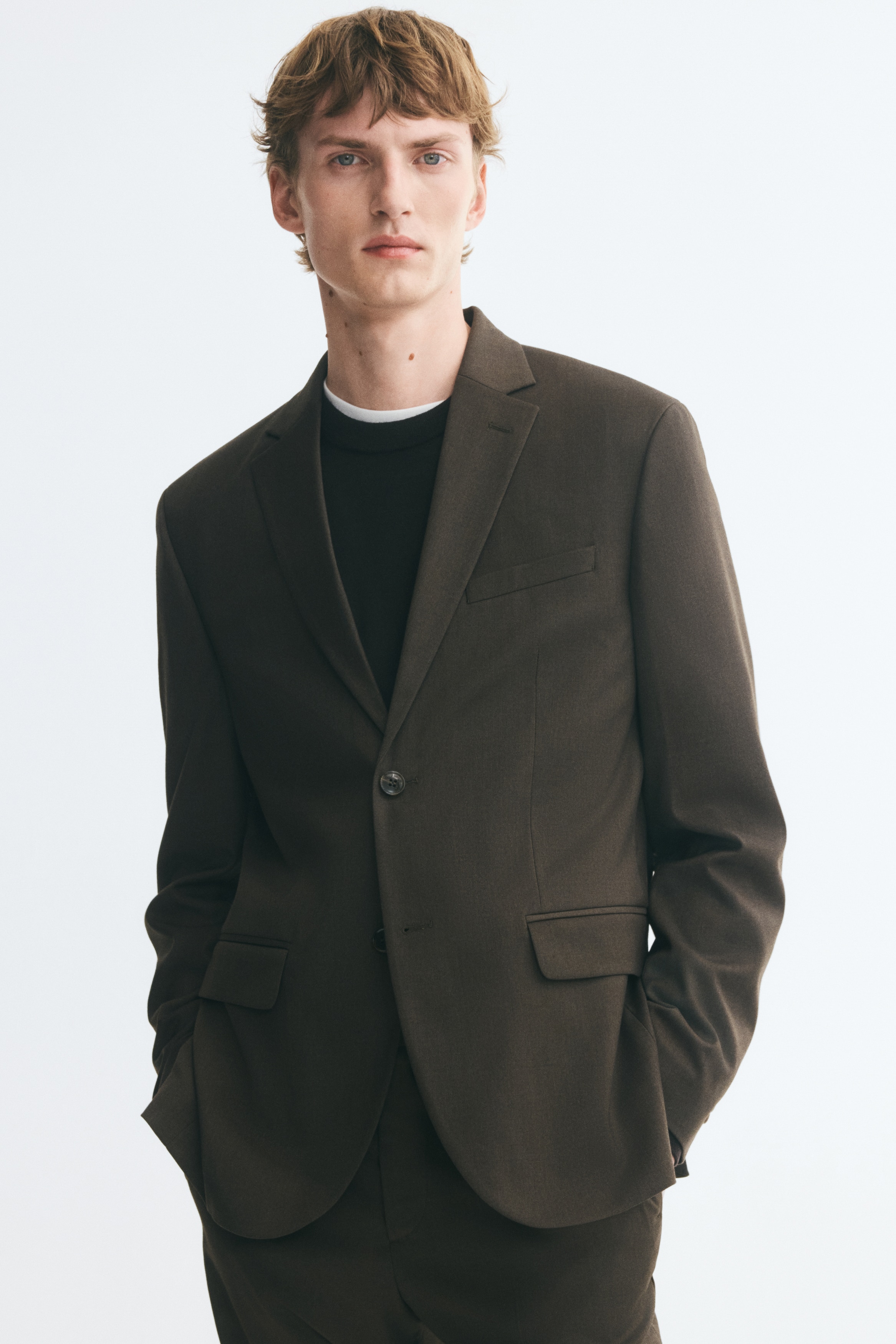 A dark brown, single-breasted suit jacket is shown front-facing, featuring notch lapels, a welt chest pocket, and two lower flap pockets. The jacket presents a structured, regular-fit silhouette with a single button fastened at the front.