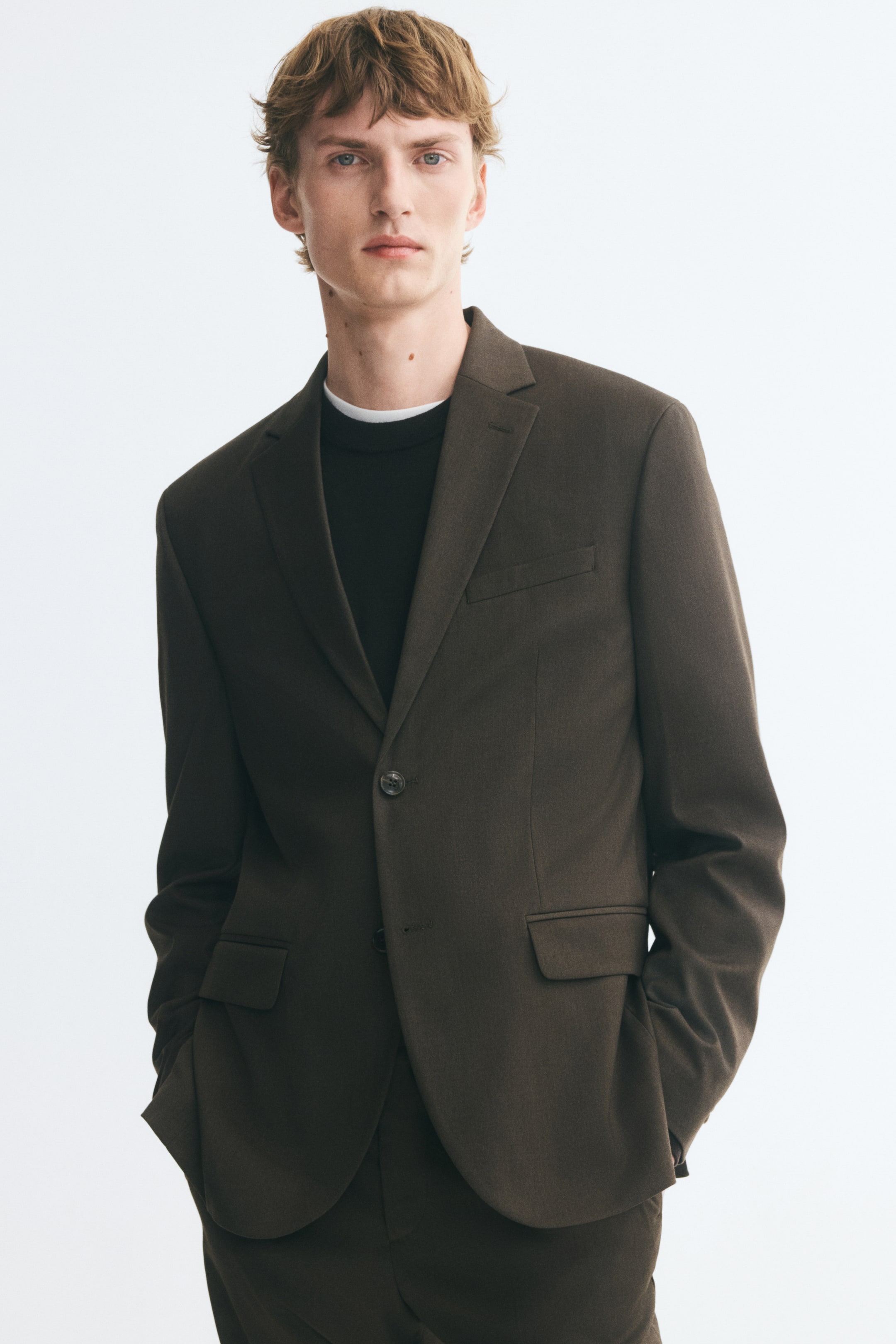 View larger image: The model is wearing a dark brown single-breasted jacket over a black crew-neck top and matching trousers. The jacket features notch lapels, a front button closure, a welt chest pocket, and flap front pockets.
