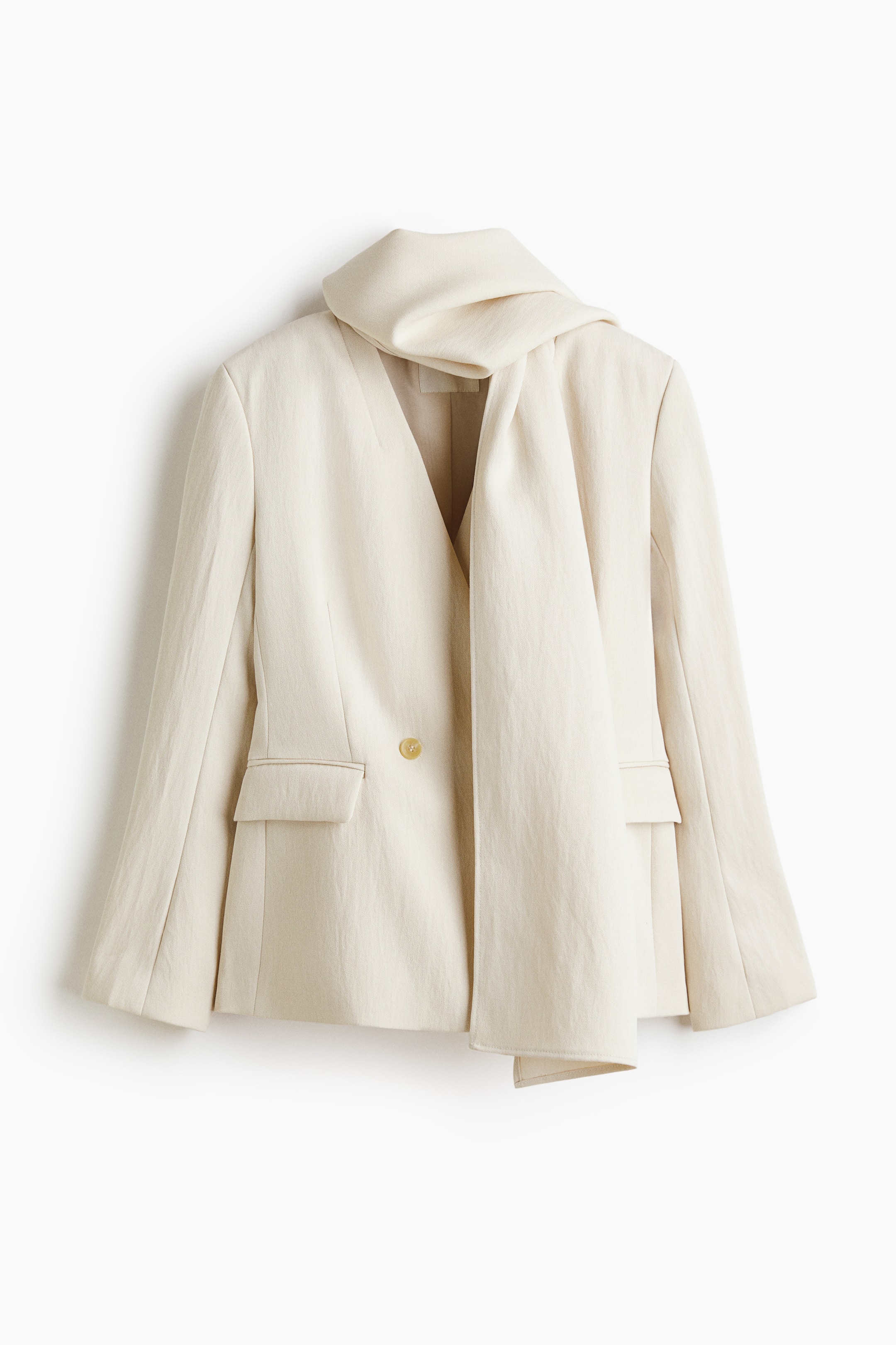 Vis større bilde: En lys beige blazer med avslappet passform viser en dyp V-utringning med et integrert drapert skjerf. Den har også to frontlommer med klaff og en enkelt lysebrun knappelukking.