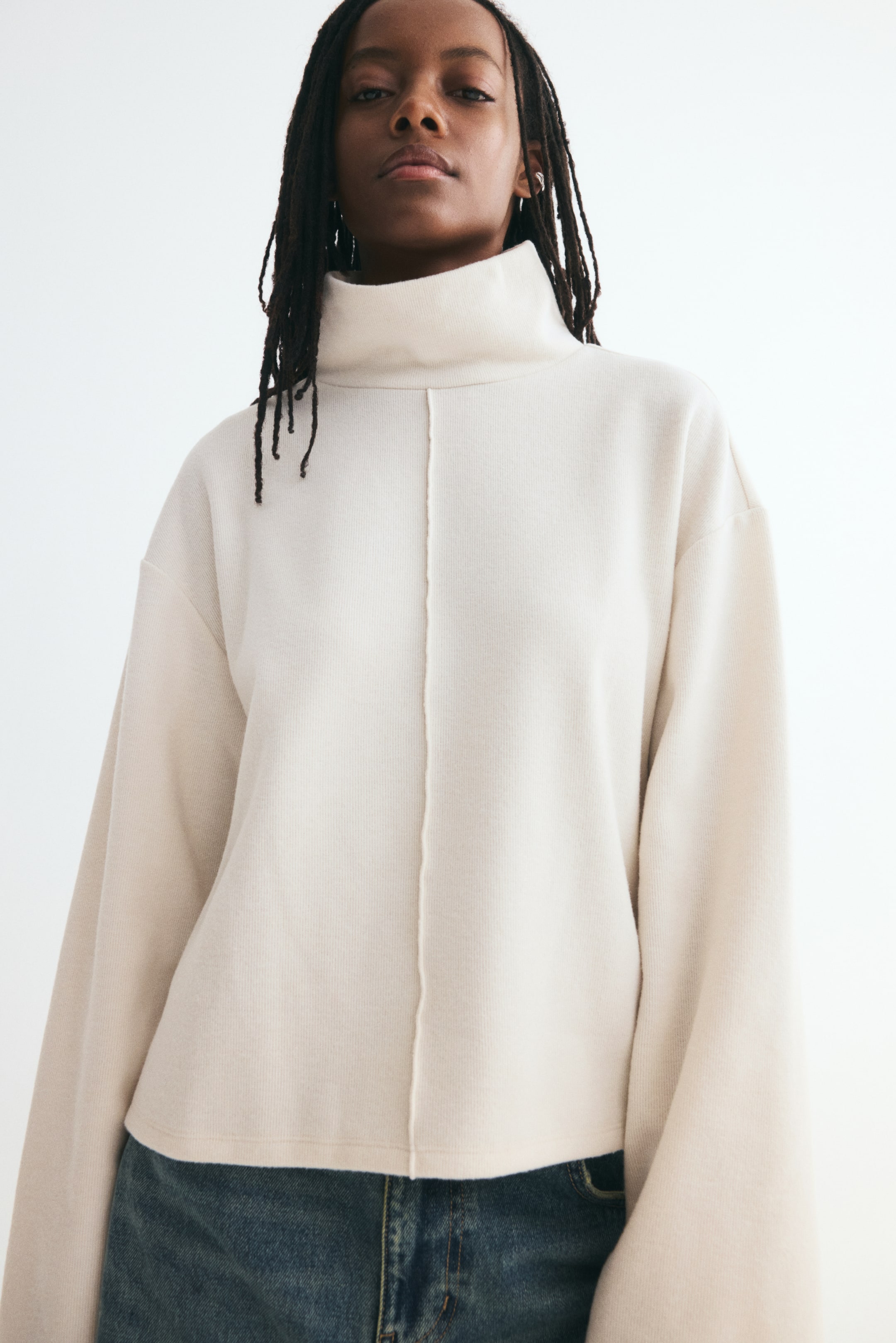 View larger image: A loose-fit, off-white knit sweater is shown from the front, featuring a high neck, dropped shoulders, and wide, long sleeves. A prominent vertical seam runs down the centre front of the sweater.