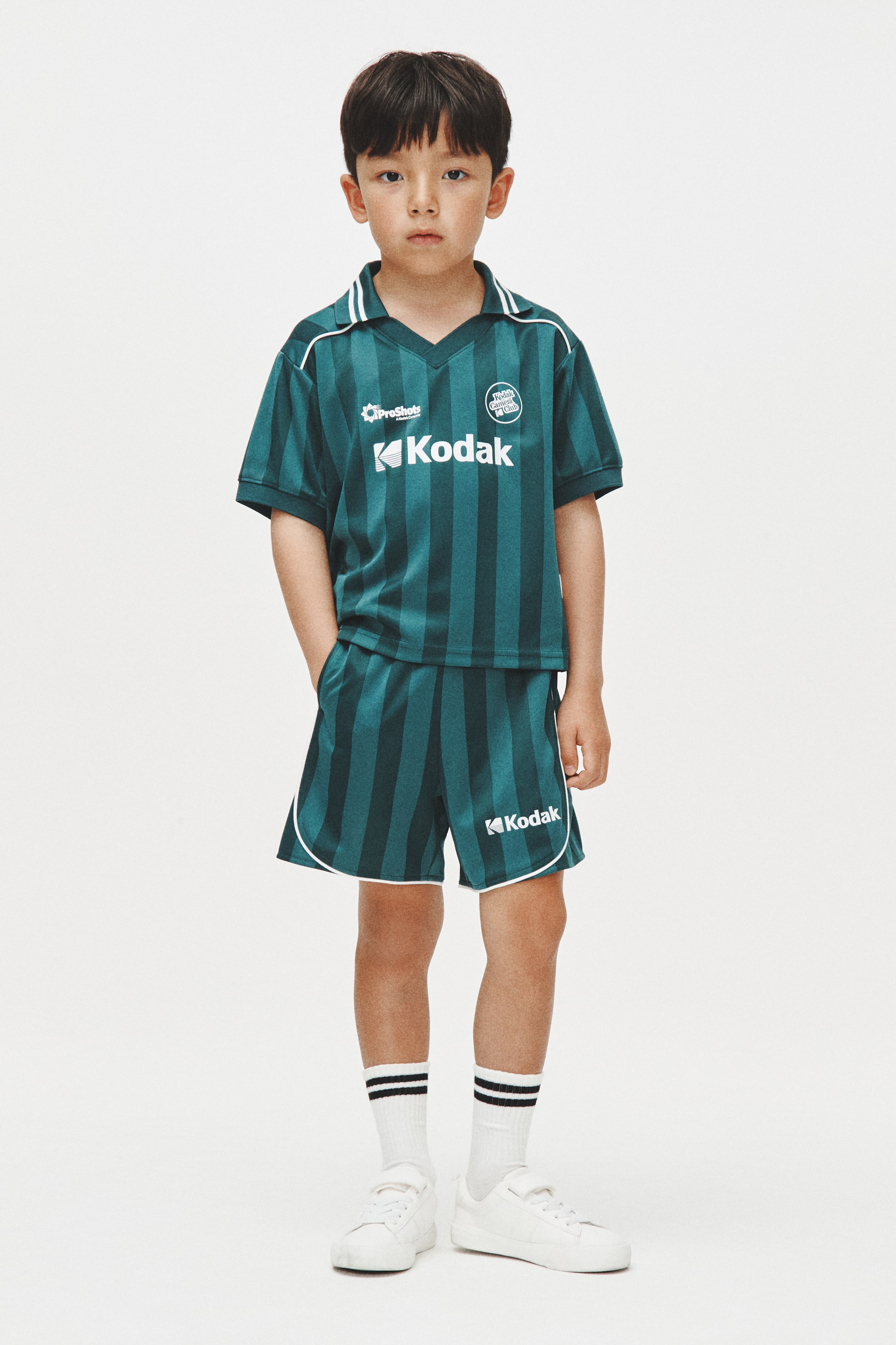 View larger image: Suot ng modelo ang isang teal at dark green na striped na short-sleeved collared shirt at shorts set. Ang shirt ay may puting piping, "ProShots" logo, at "Kodak" logo, habang ang shorts ay may puting piping sa hem at "Kodak" logo. Nakatayo ang modelo na may isang kamay sa bulsa ng shorts.
