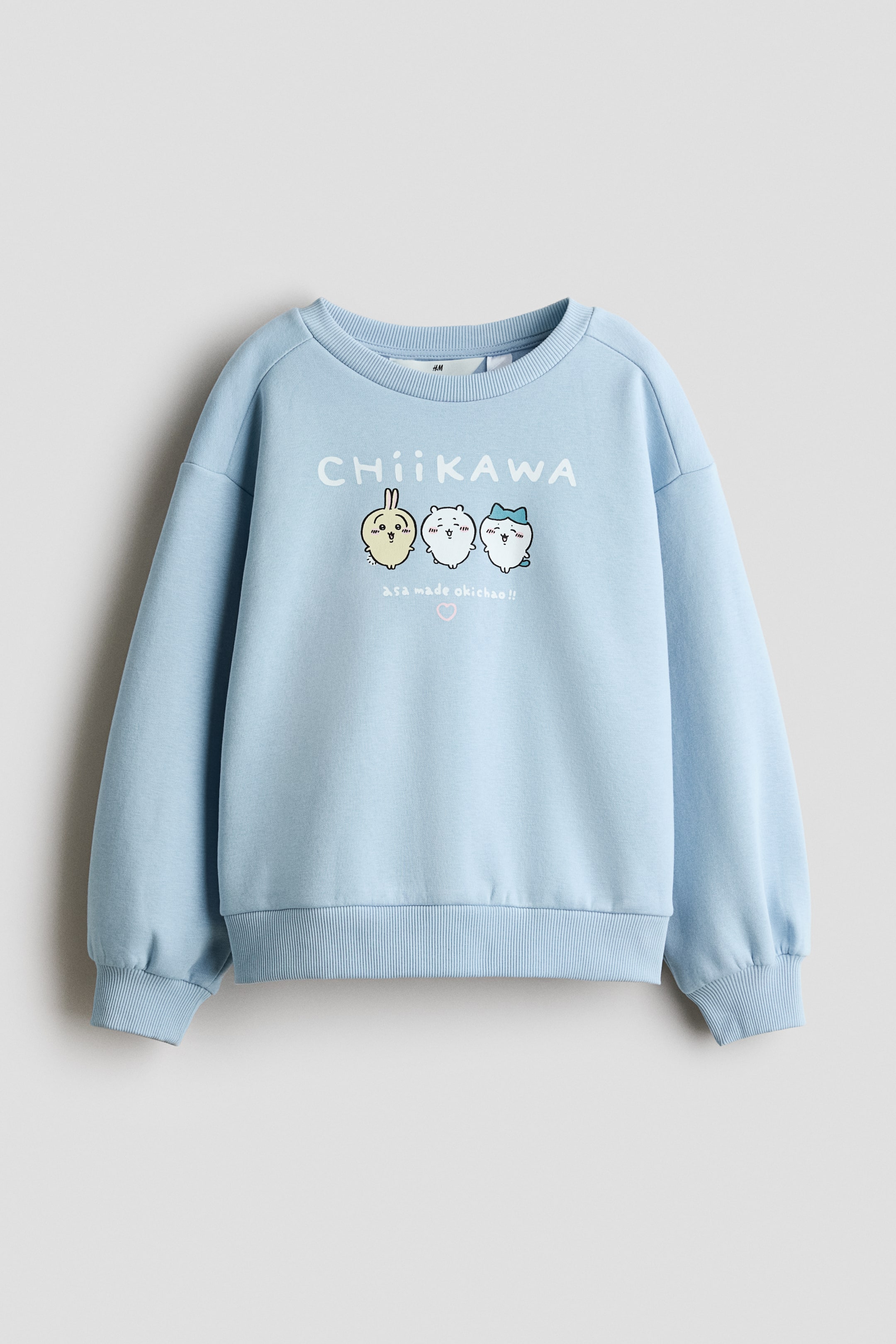 Ver imagem maior: Uma camisola de felpa azul-clara está vista de frente, exibindo uma gola redonda com acabamento canelado, ombros descaídos e punhos e bainha canelados. A parte frontal apresenta uma impressão gráfica de três personagens de desenho animado sob o texto branco "CHIIKAWA".