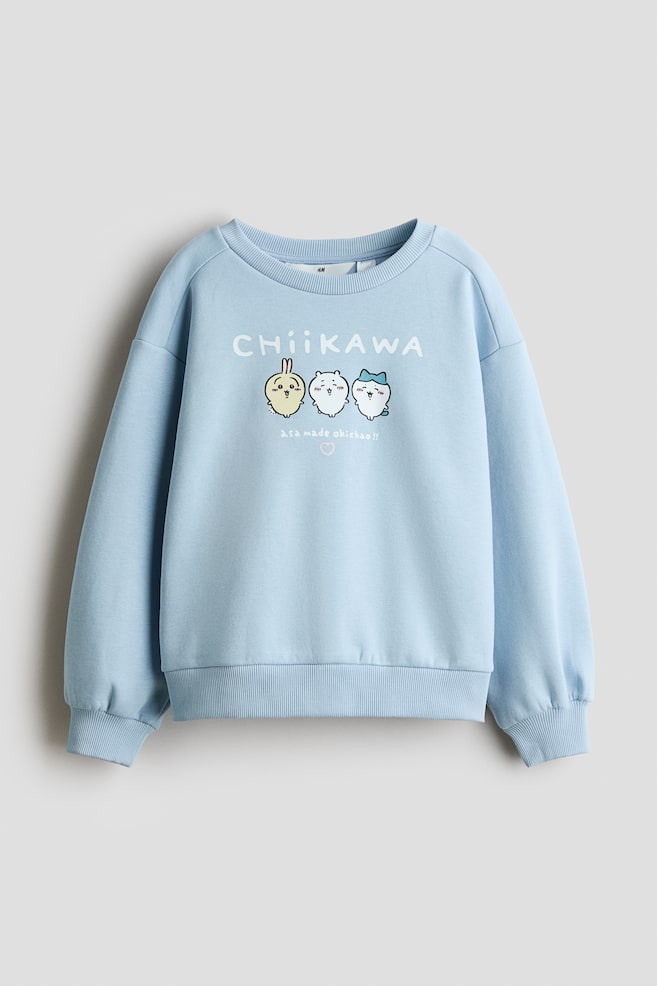 A light blue fleece jumper is shown from the front, featuring a round, rib-trimmed neckline, dropped shoulders, and ribbed cuffs and hem. The front displays a graphic print of three cartoon characters beneath the white text "CHIIKAWA". - 1
