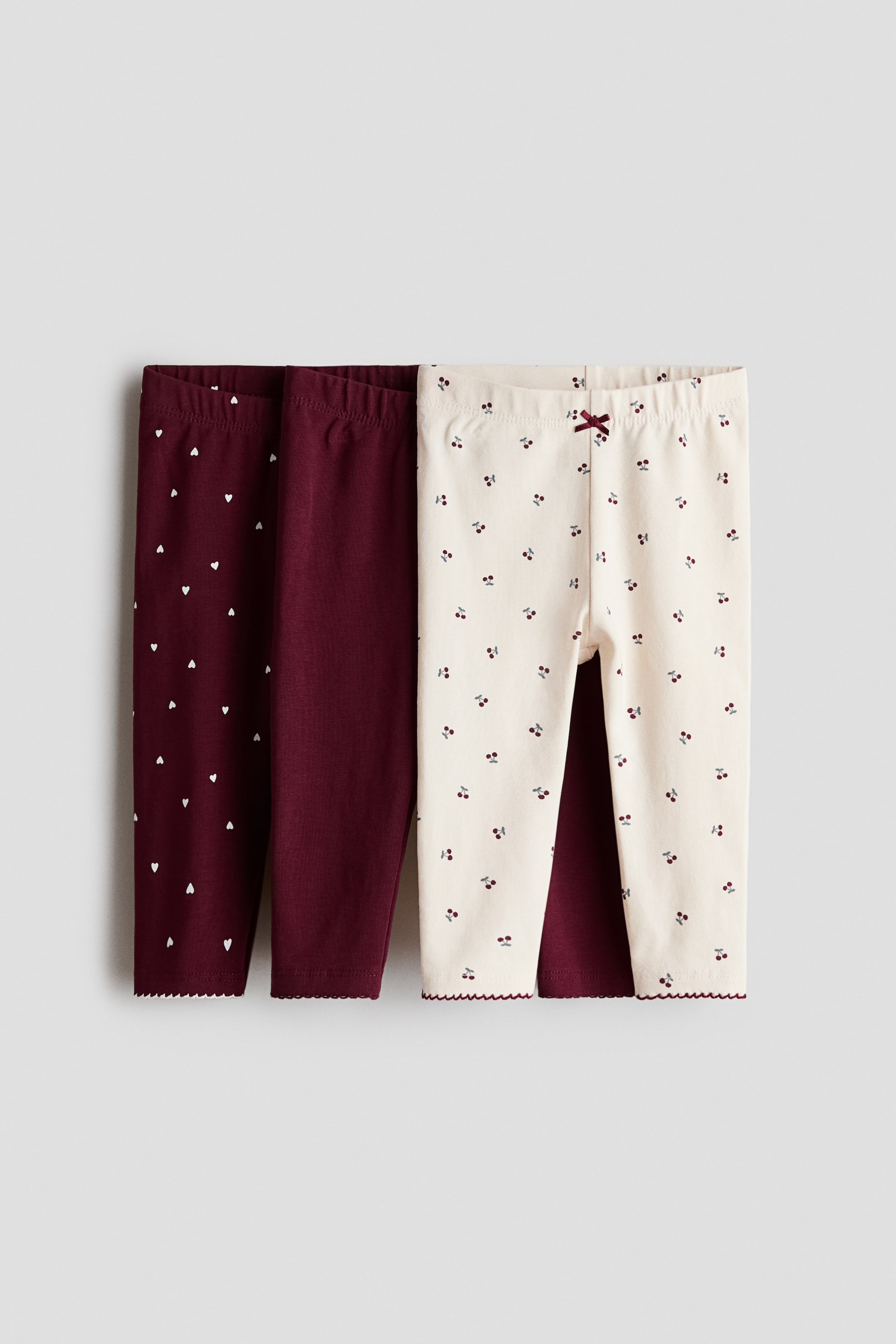 View larger image: A three-pack of children's leggings is displayed flat and front-facing, showcasing three distinct styles with elasticized waistbands and scalloped ankle hems. The set includes a burgundy pair with a small white heart pattern, a solid burgundy pair, and an off-white pair featuring a red cherry pattern with a small bow detail at the waist.