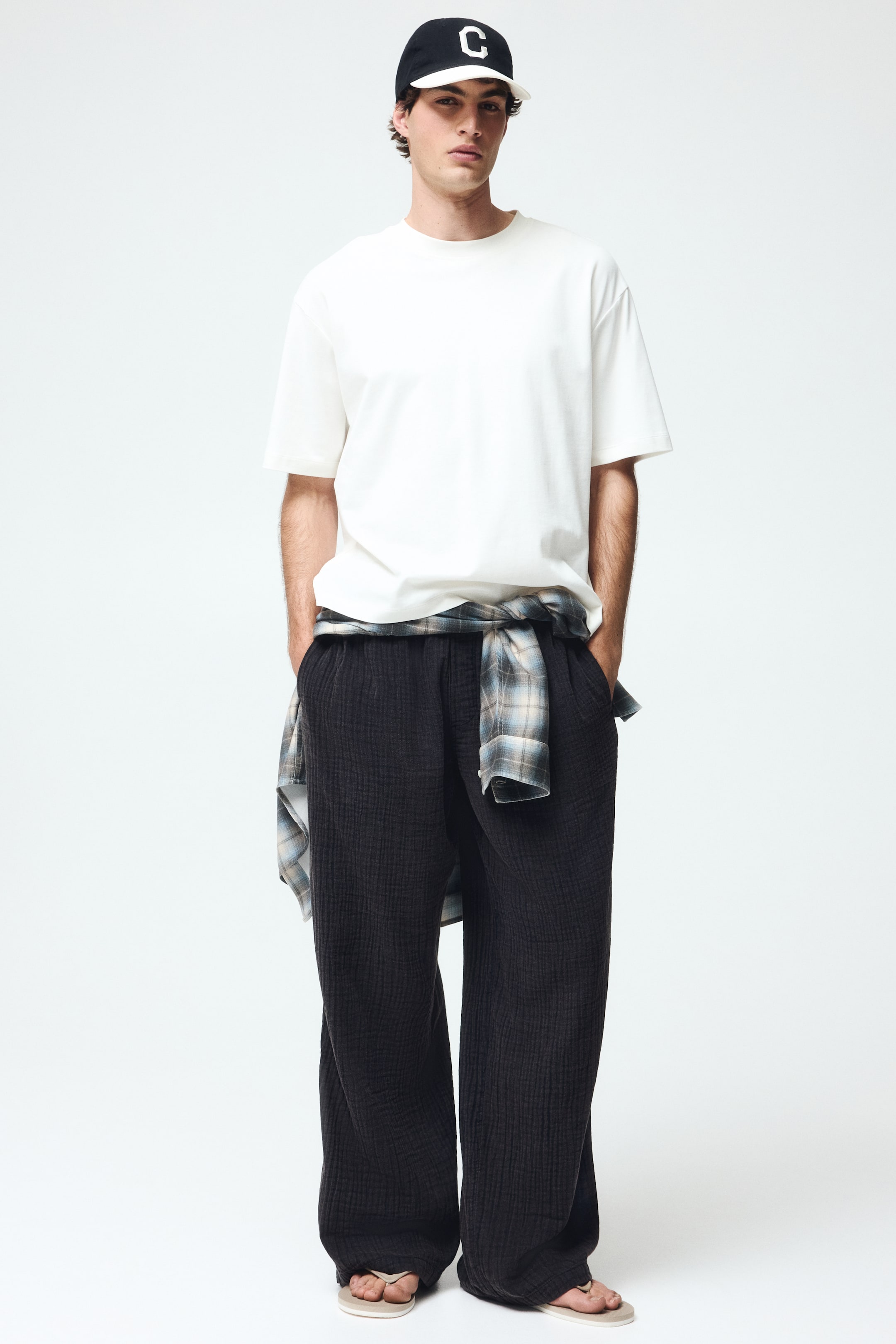 View larger image: "Ang mga itim na wide-leg trousers ay may crinkled textured fabric at maluwang na full-length fit, habang ang plaid na shirt ay nakatali sa kanilang waistband."