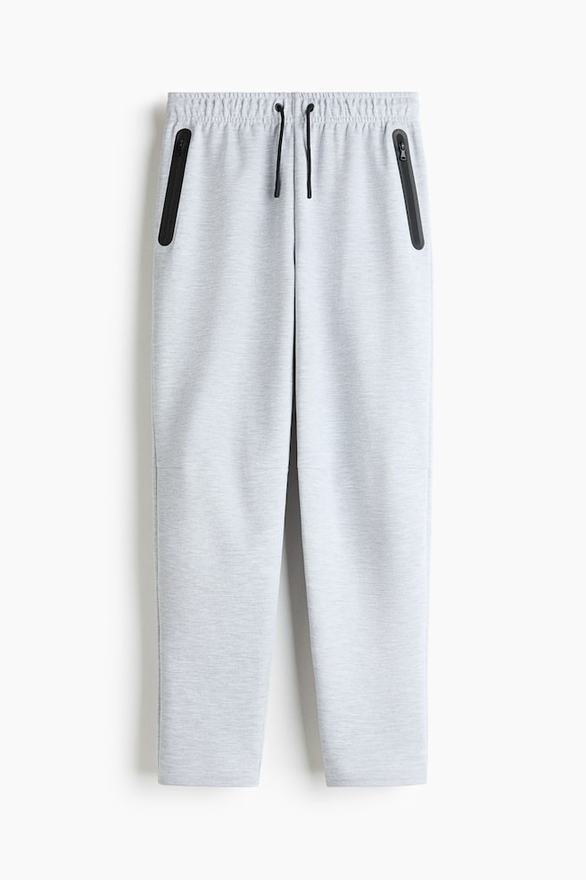 These light gray heathered sweatpants are facing front, featuring an elastic waistband with an adjustable black drawstring. Two vertical side pockets with black-trimmed zippers are detailing the upper hips, while the legs are tapering slightly toward the bottom. - 1