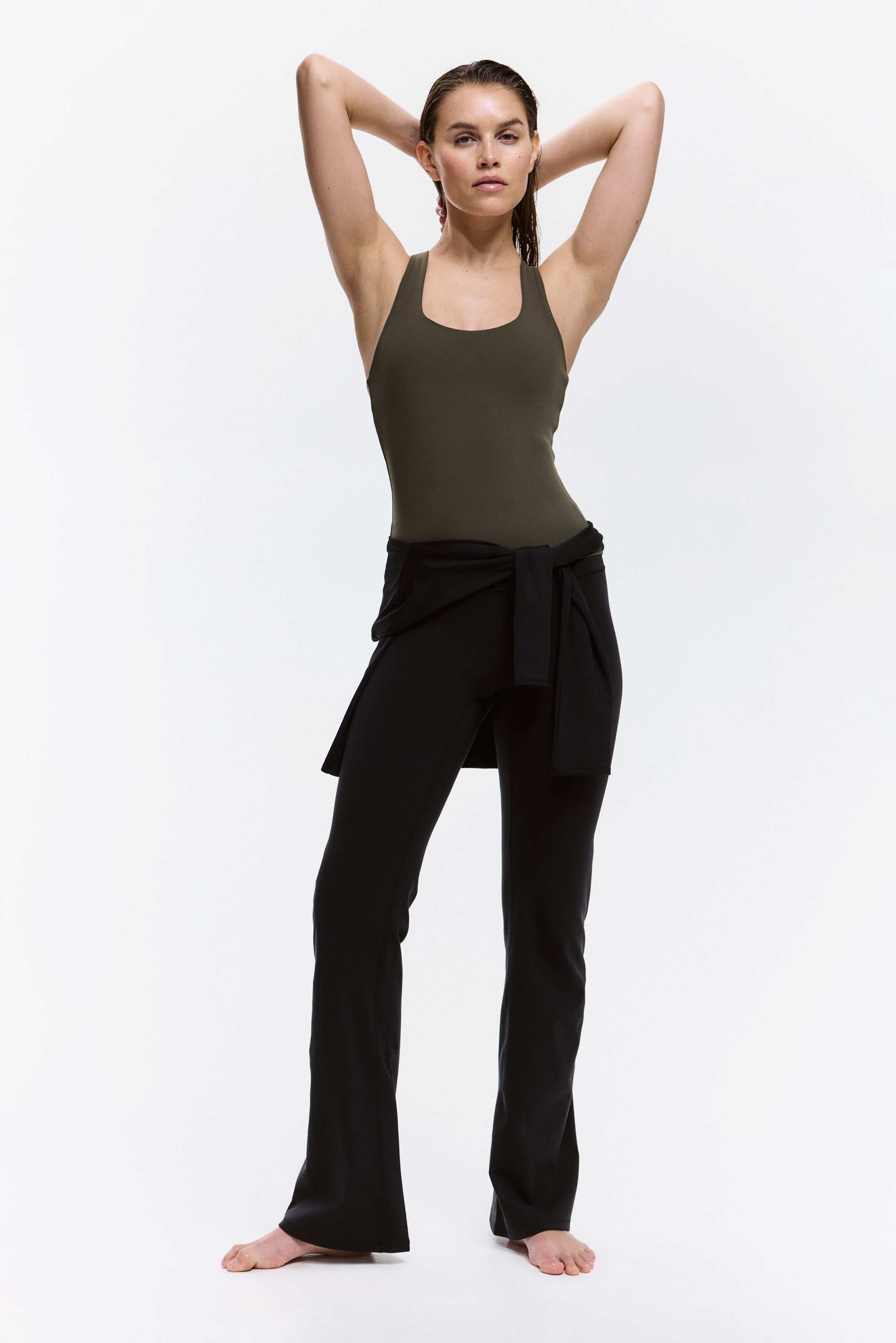 Ingrandisci l'immagine: Un top sportivo verde oliva con reggiseno integrato e tecnologia SoftMove?, caratterizzato da uno scollo a barchetta e spalline larghe, indossato dalla modella. La modella è in piedi con le braccia sollevate sopra la testa, mettendo in risalto la vestibilità aderente del top, mentre indossa pantaloni neri e un indumento nero è legato intorno alla vita.