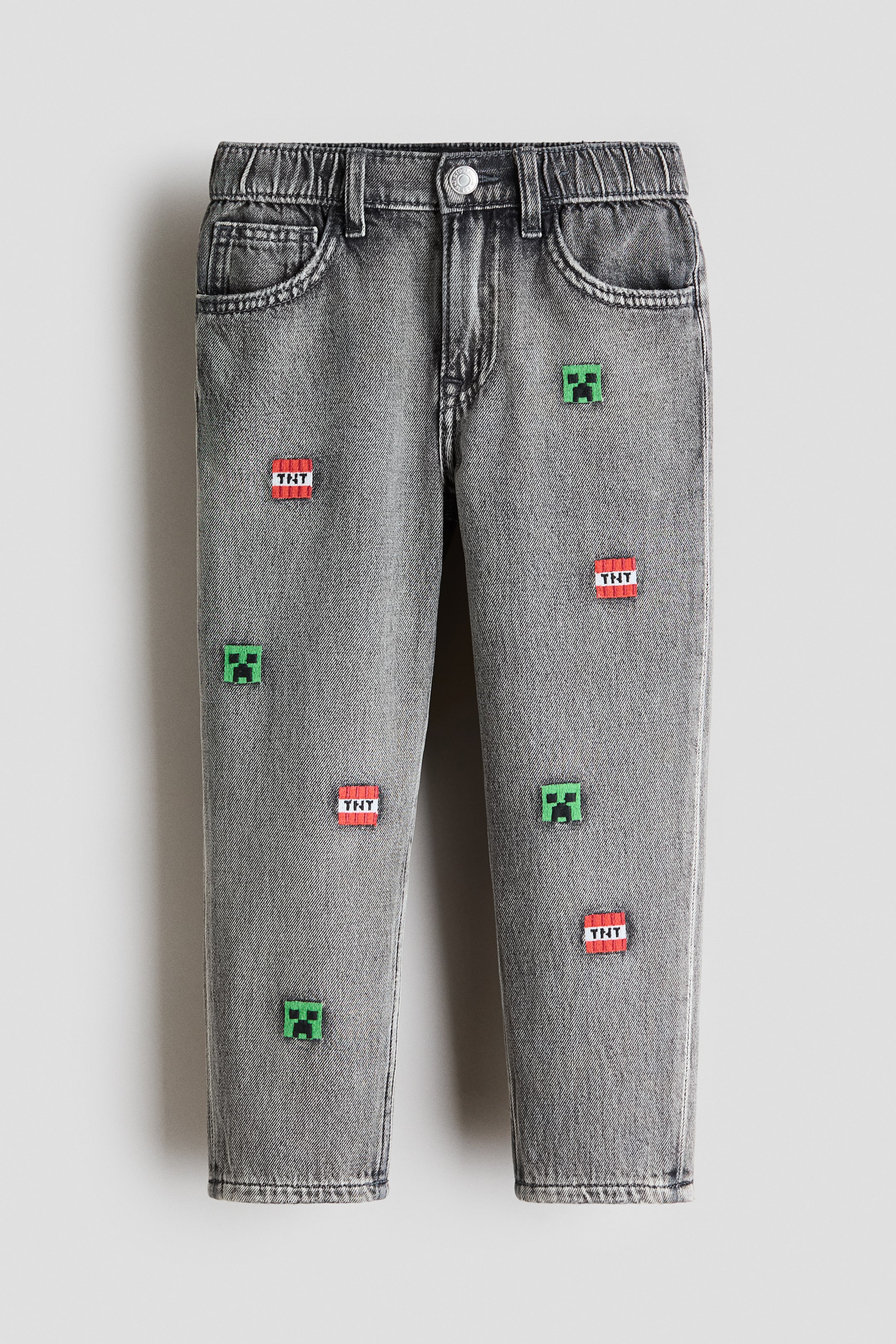 View larger image: Grey denim jeans featuring an elasticated back waistband, a front button closure, and scoop pockets. The straight-leg jeans display an all-over pattern of embroidered pixelated green Creeper faces and red TNT blocks.