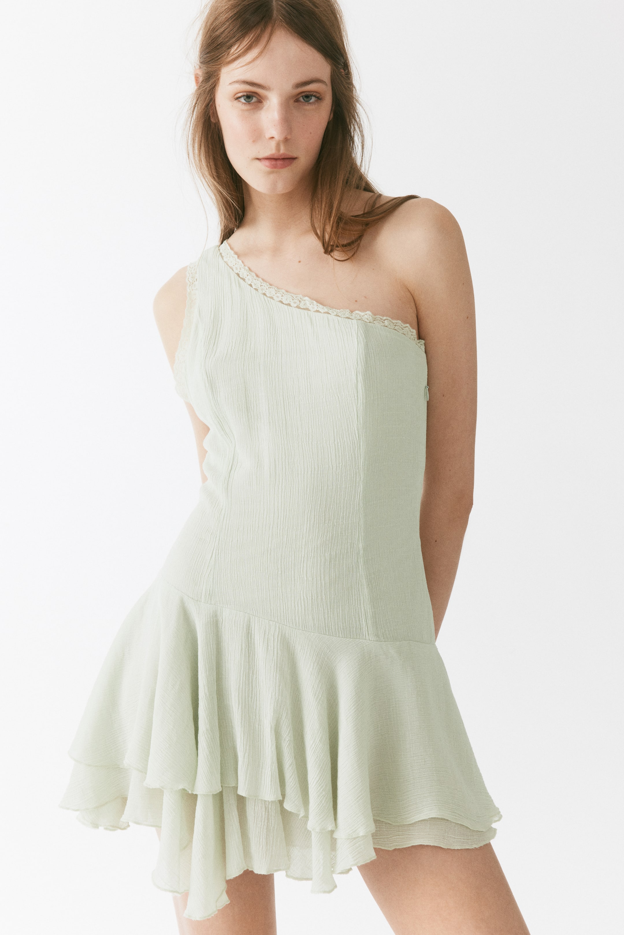 View larger image: A light green one-shoulder dress featuring a crinkle-textured fabric and delicate lace trim along the single shoulder strap, with tiered ruffles creating an asymmetrical hemline.