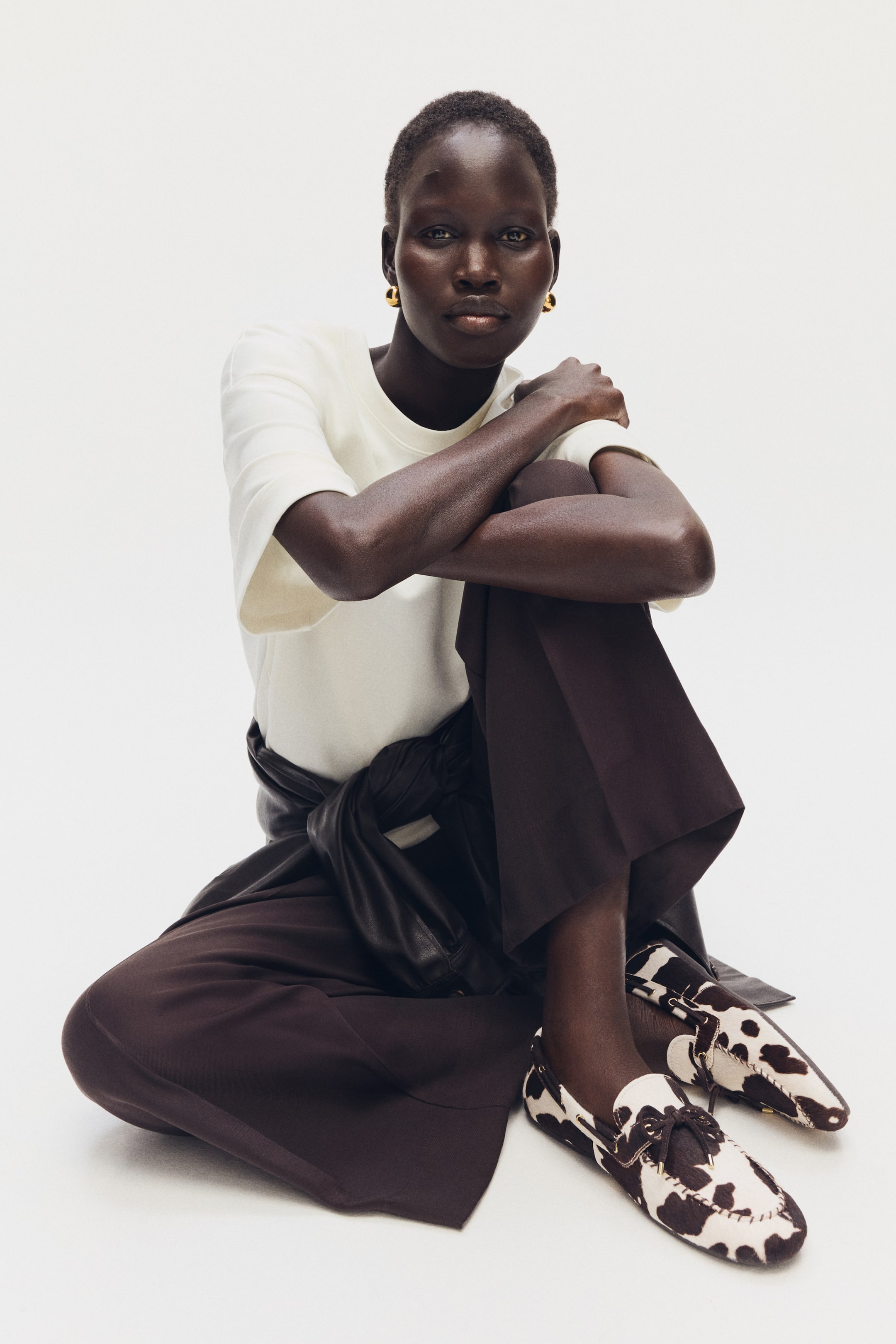 Преглед на по-голямо изображение: Кафяво-бели мокасини с принт на крава, които се отличават с текстурирана горна част с шевове тип мокасин и детайл с връзки, който образува панделка на горната част. Те са представени в преден и леко страничен изглед.